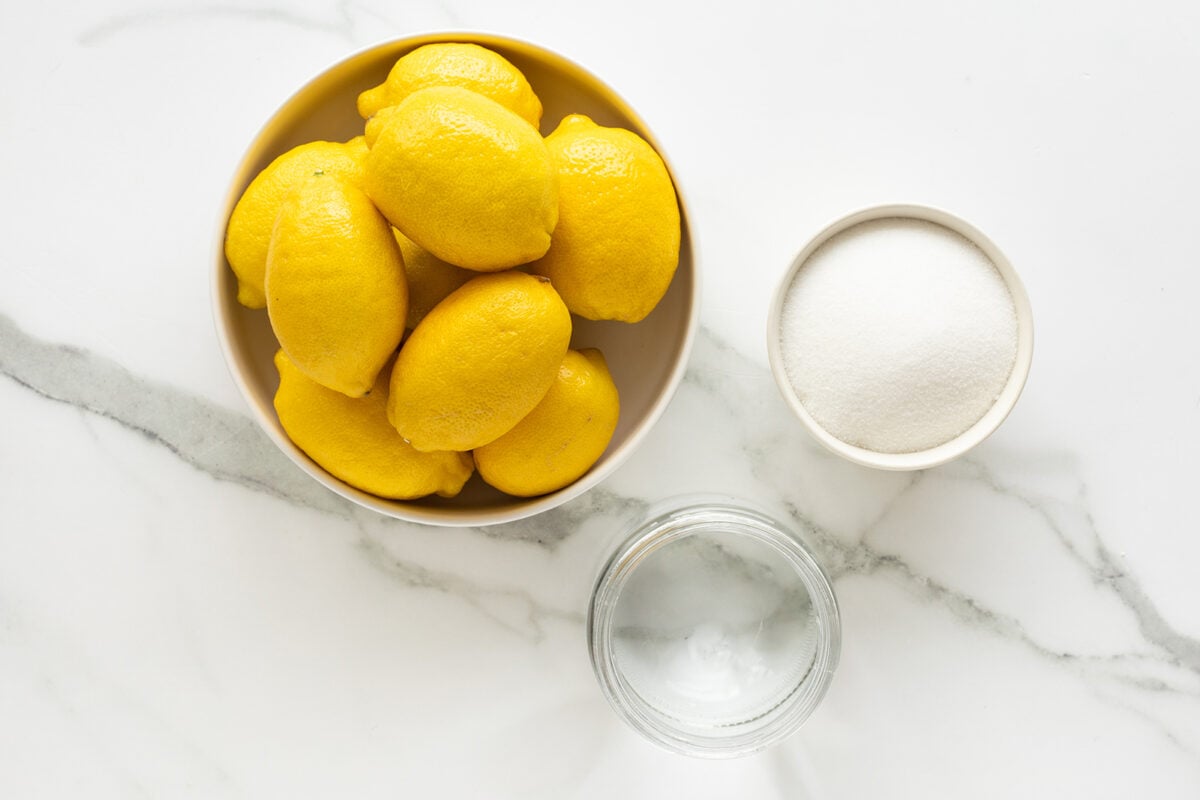 A bowl of whole yellow lemons, a small bowl of white sugar, and a glass of water are arranged on a white marble surface.