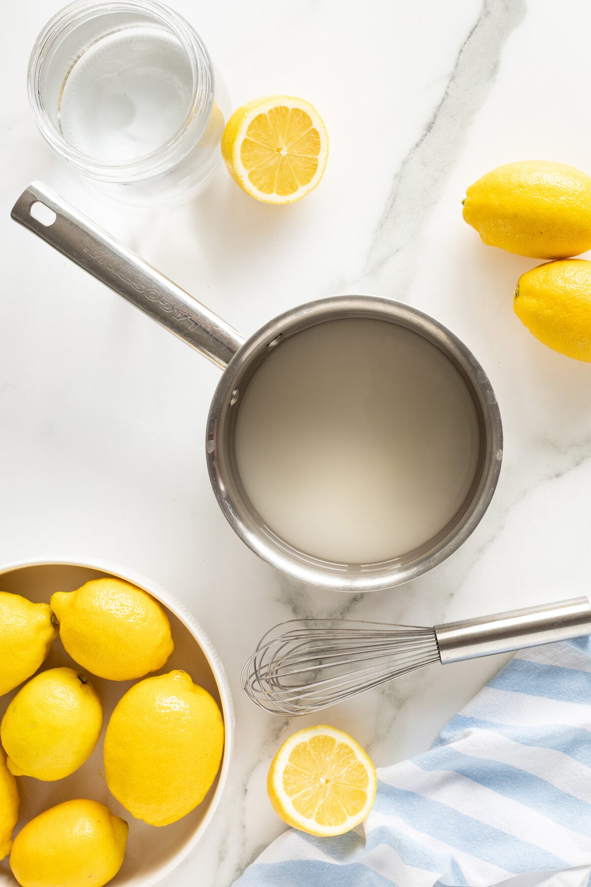 A saucepan with a cloudy liquid sits on a marble surface, surrounded by fresh lemons, a bowl of lemons, a glass of water, a whisk, and a blue-striped cloth.