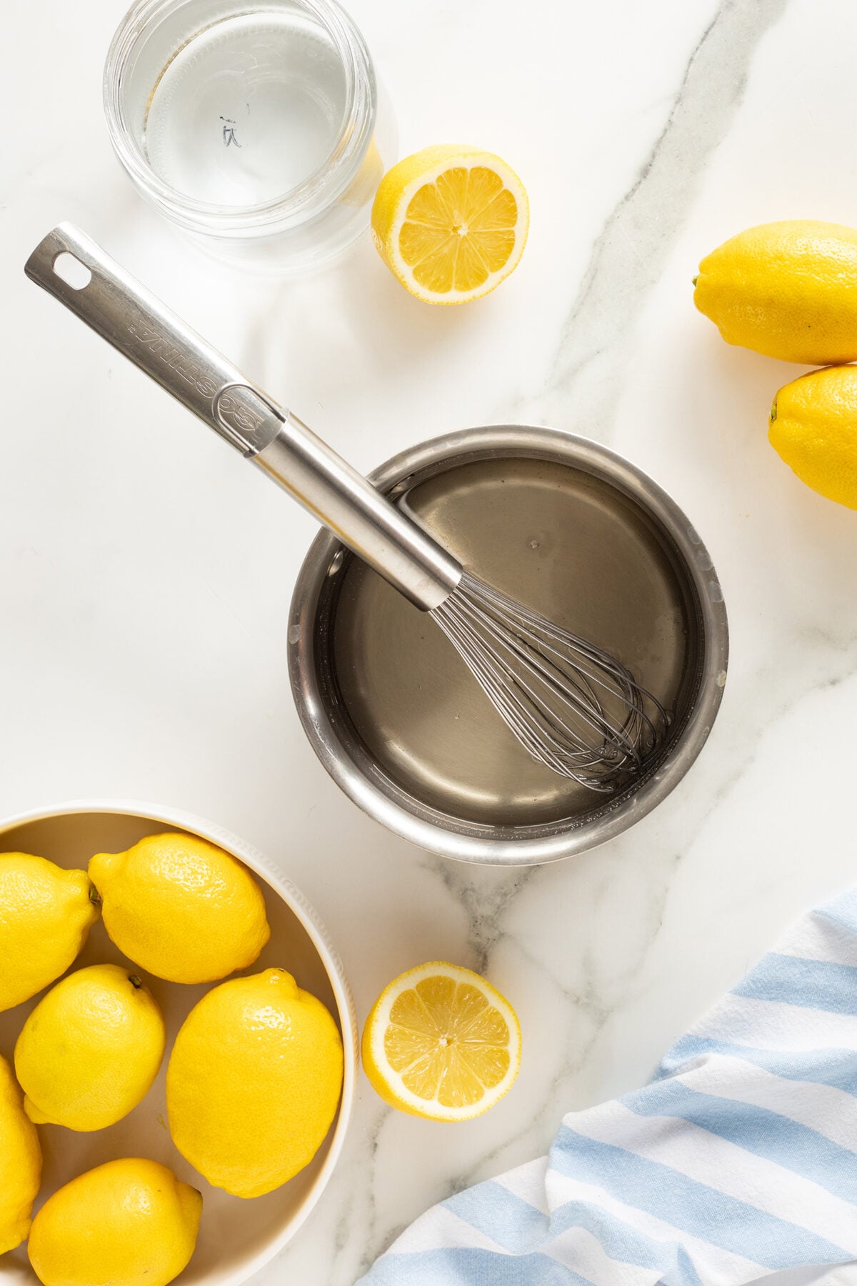 A metal whisk in a bowl with liquid, surrounded by fresh lemons, a halved lemon, a glass of water, and a striped cloth on a white marble surface.