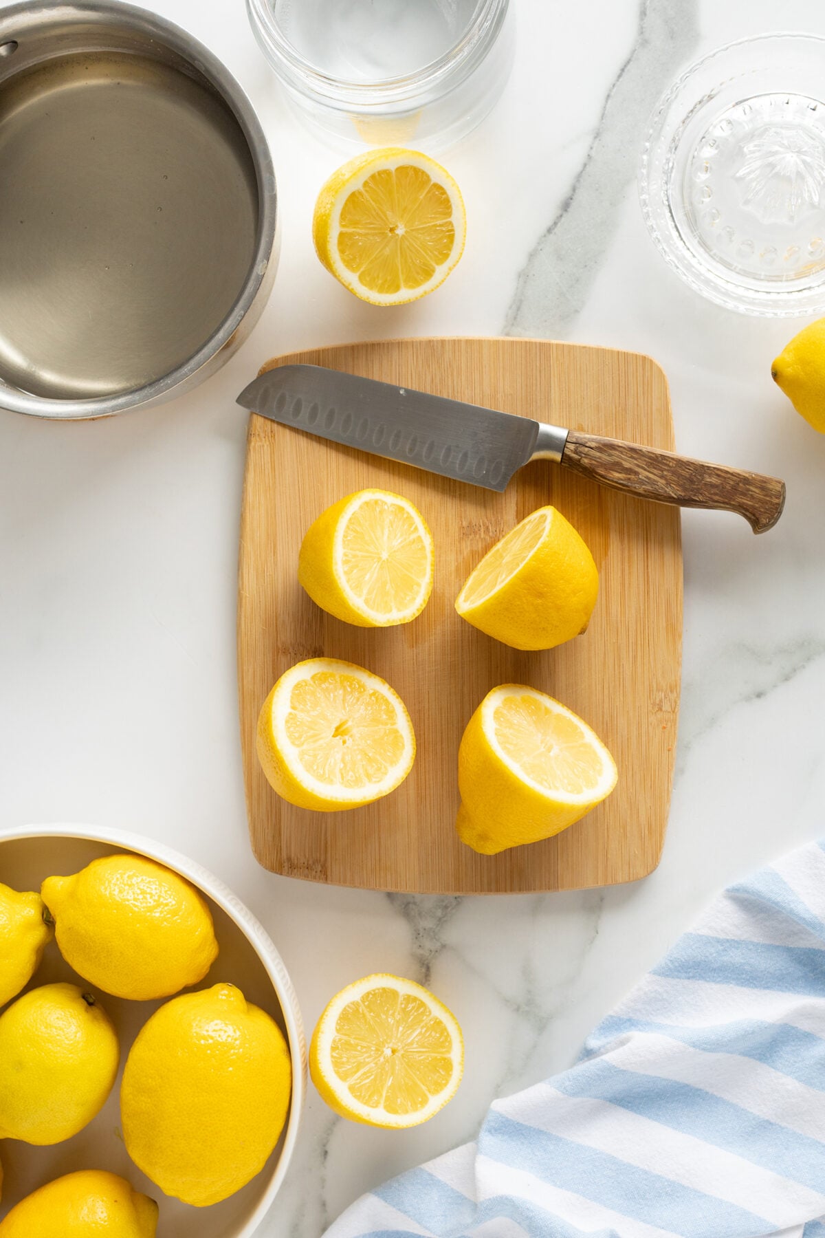 A wooden cutting board with a knife and halved lemons sits on a marble counter. Whole lemons, a bowl of water, a striped towel, and a glass citrus juicer are nearby.