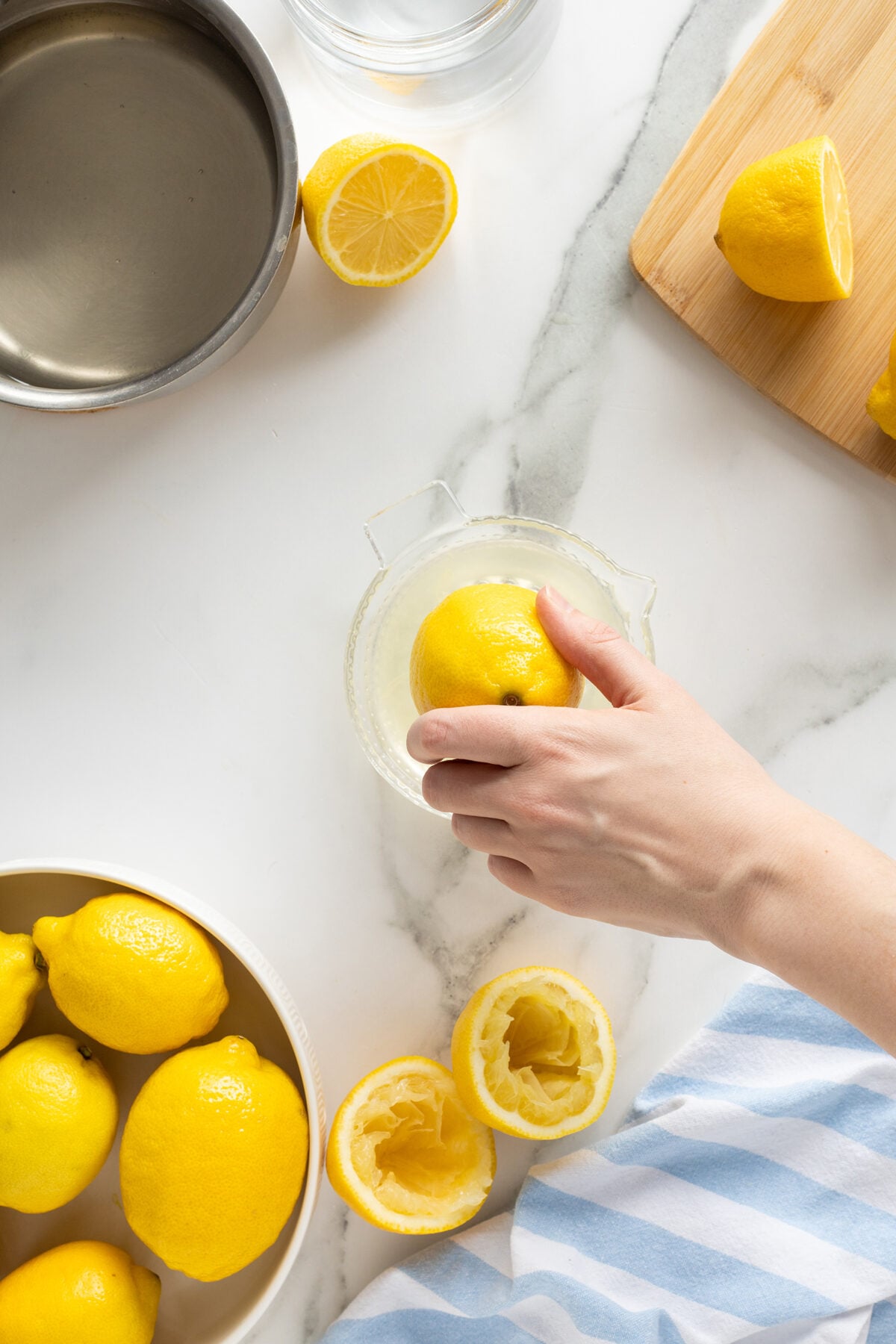 A hand squeezes a lemon on a glass juicer surrounded by whole and halved lemons, a bowl of lemons, a pot of water, and a striped cloth on a white marble countertop.
