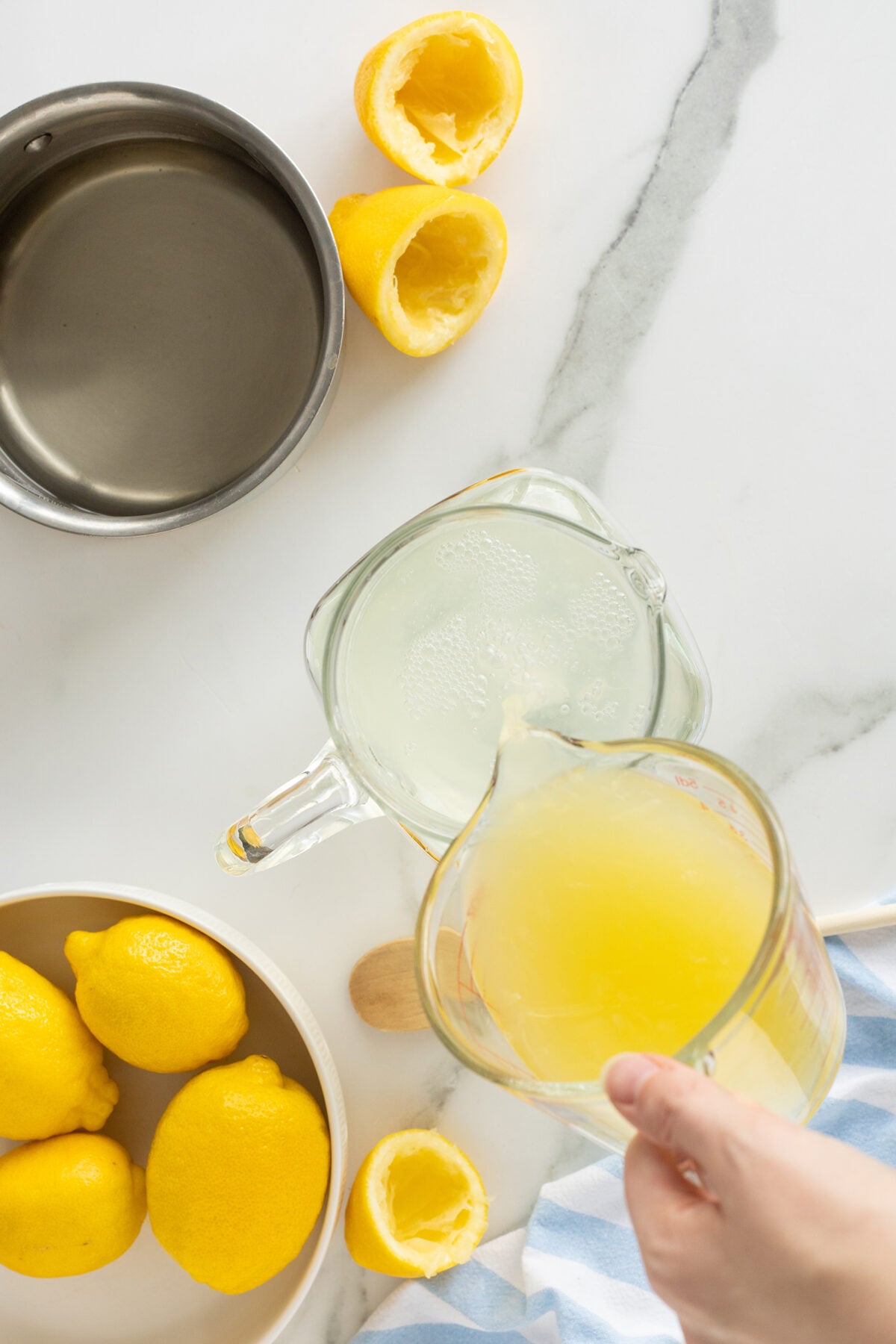 A hand pours fresh lemon juice from a measuring cup into a glass container. Nearby are squeezed lemon halves, whole lemons in a bowl, a saucepan with water, and a blue striped cloth on a marble surface.