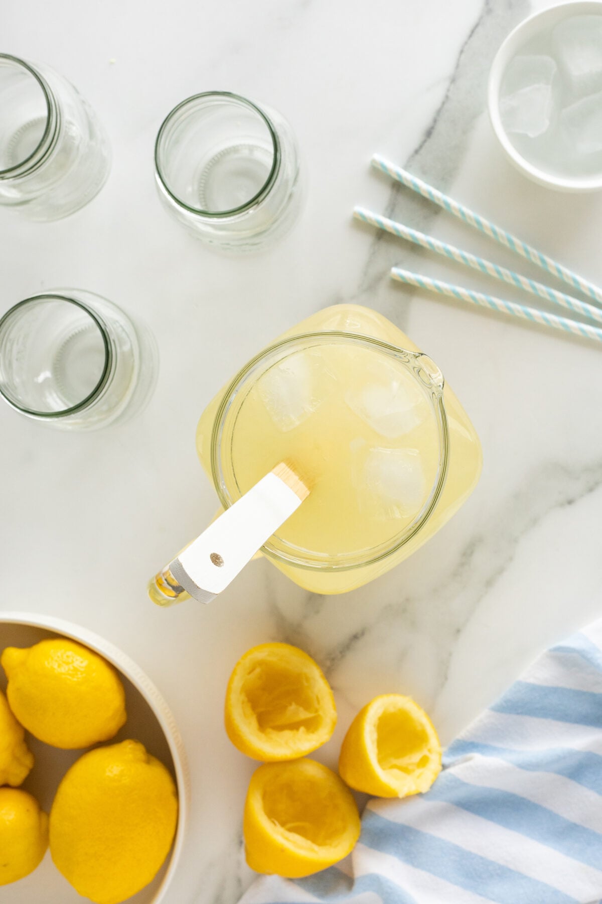 A pitcher of lemonade with ice sits on a marble surface, surrounded by empty glass jars, blue-striped straws, ice cubes, whole and squeezed lemons, and a blue-striped cloth.