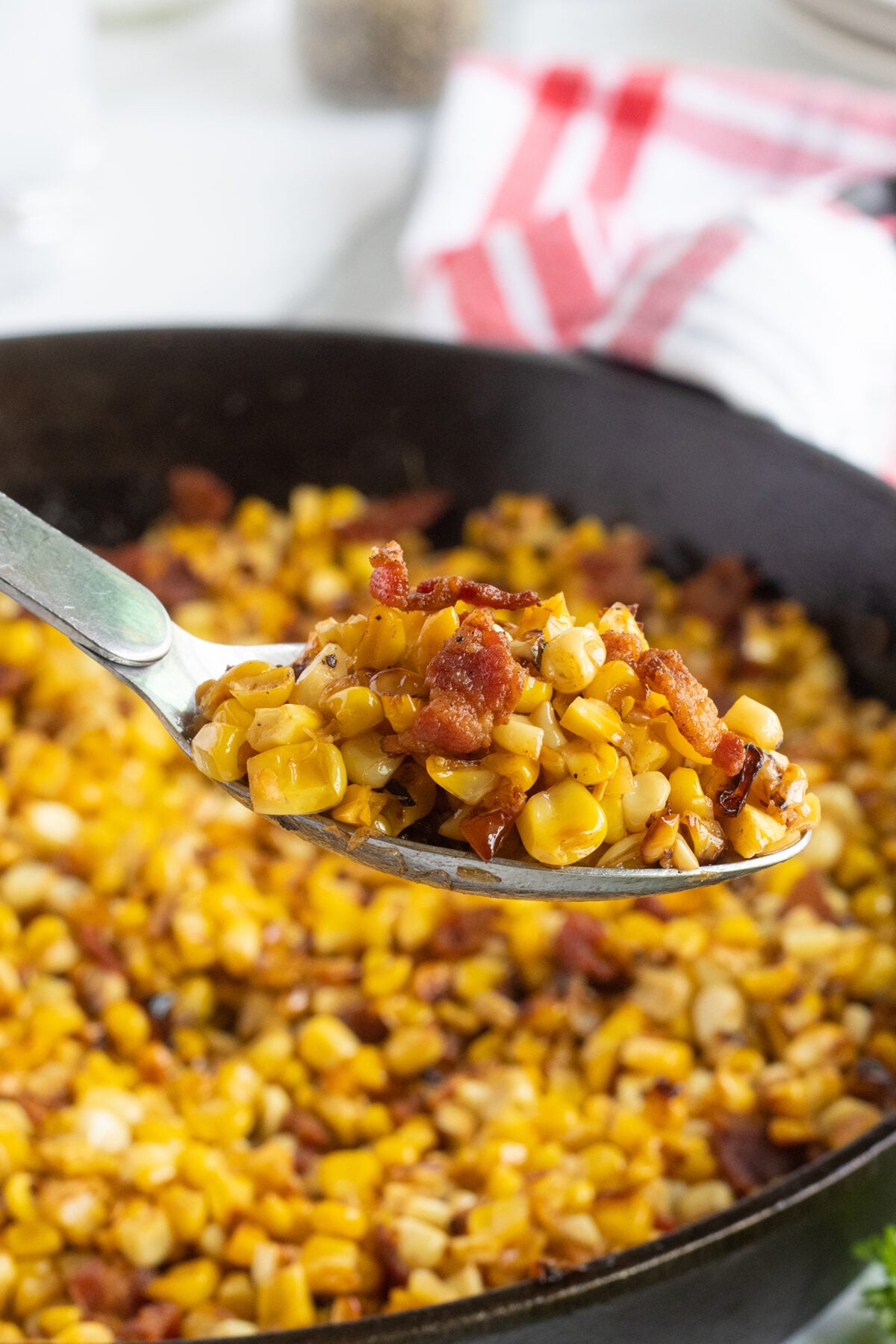 A close-up of a spoon holding sautéed corn mixed with crispy bacon bits, with more of the same mixture in a skillet in the background. A red and white towel sits in the blurred background.