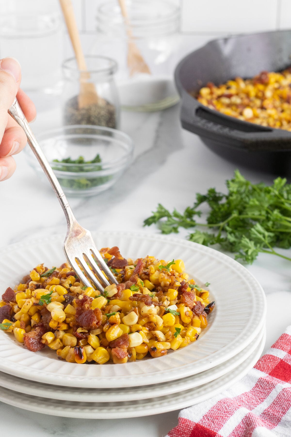 A hand holds a fork over a plate of sautéed corn mixed with bacon and herbs, with more corn in a skillet and fresh parsley in the background on a white kitchen counter.