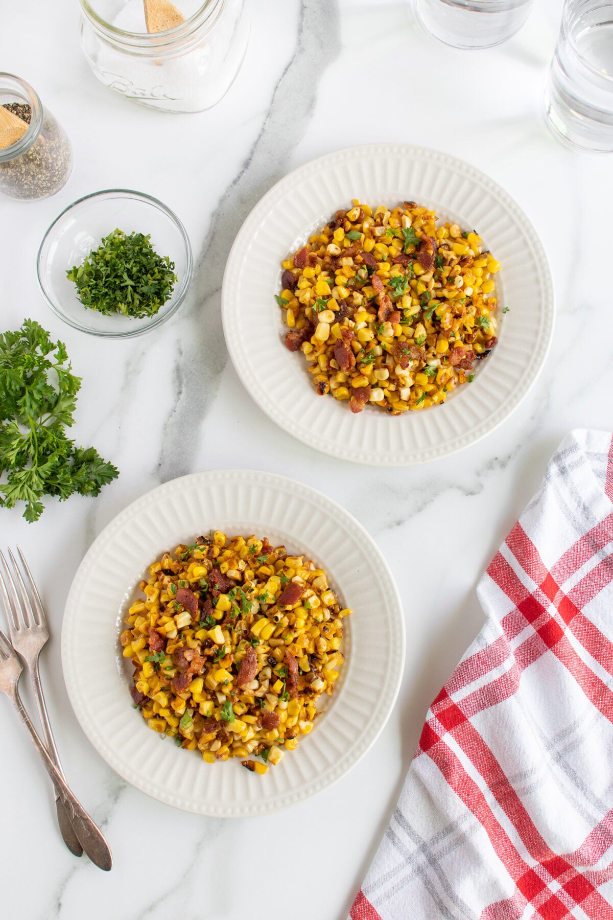 Two white plates filled with a corn, bean, and herb salad sit on a marble surface. Nearby are forks, fresh parsley, a bowl of chopped herbs, and a red-and-white checkered towel.