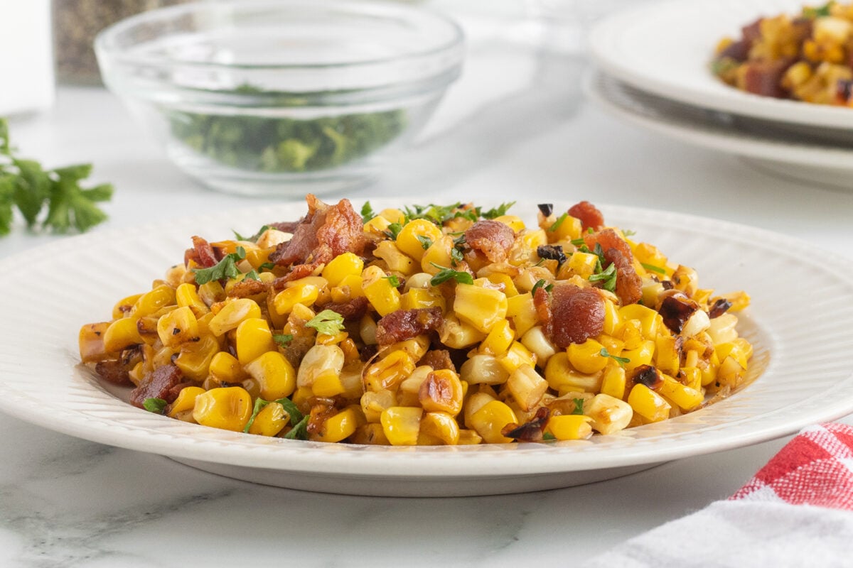 A white plate filled with a cooked corn dish mixed with chopped bacon and herbs, set on a white table with a glass bowl of parsley and another plate of food in the background.