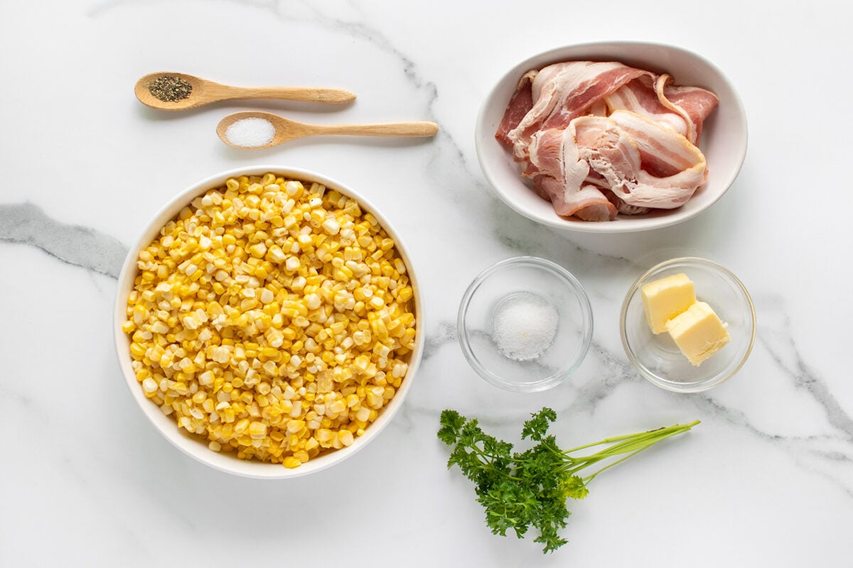 Overhead view of ingredients on a white surface: a bowl of corn kernels, a dish with raw bacon strips, butter, salt, pepper, and parsley, each in separate bowls or on spoons.