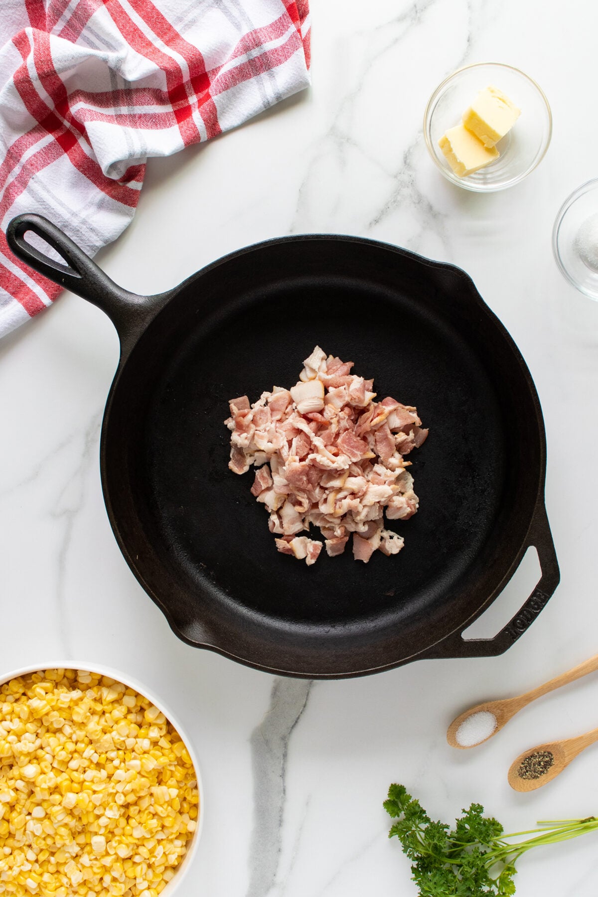 A cast iron skillet with chopped raw bacon in the center. Surrounding the skillet are a bowl of corn kernels, a pat of butter, salt, pepper, parsley, a wooden spoon, and a red-striped towel on a marble surface.
