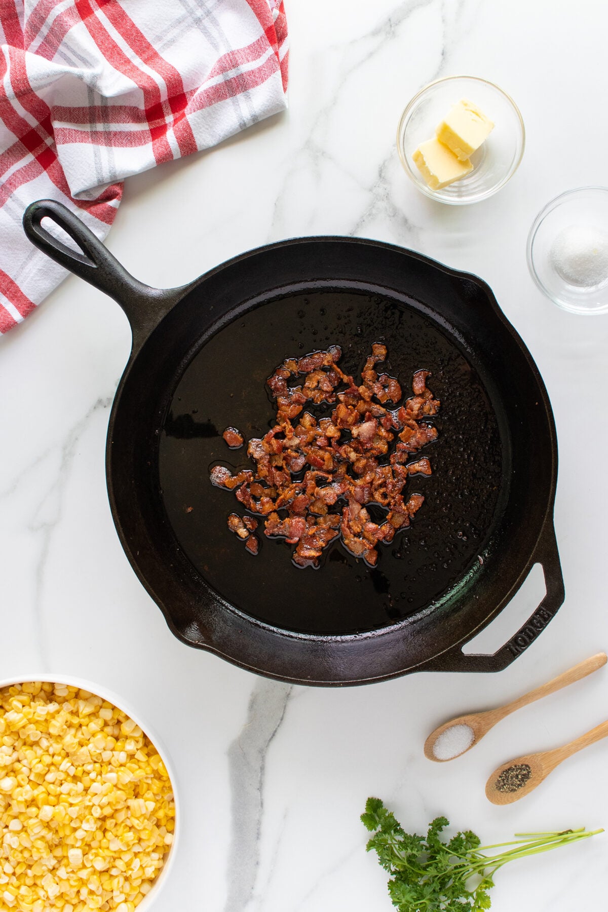 A cast iron skillet with cooked bacon pieces sits on a marble surface. Surrounding the skillet are bowls of corn, butter, salt, pepper, fresh cilantro, and a red-striped towel.