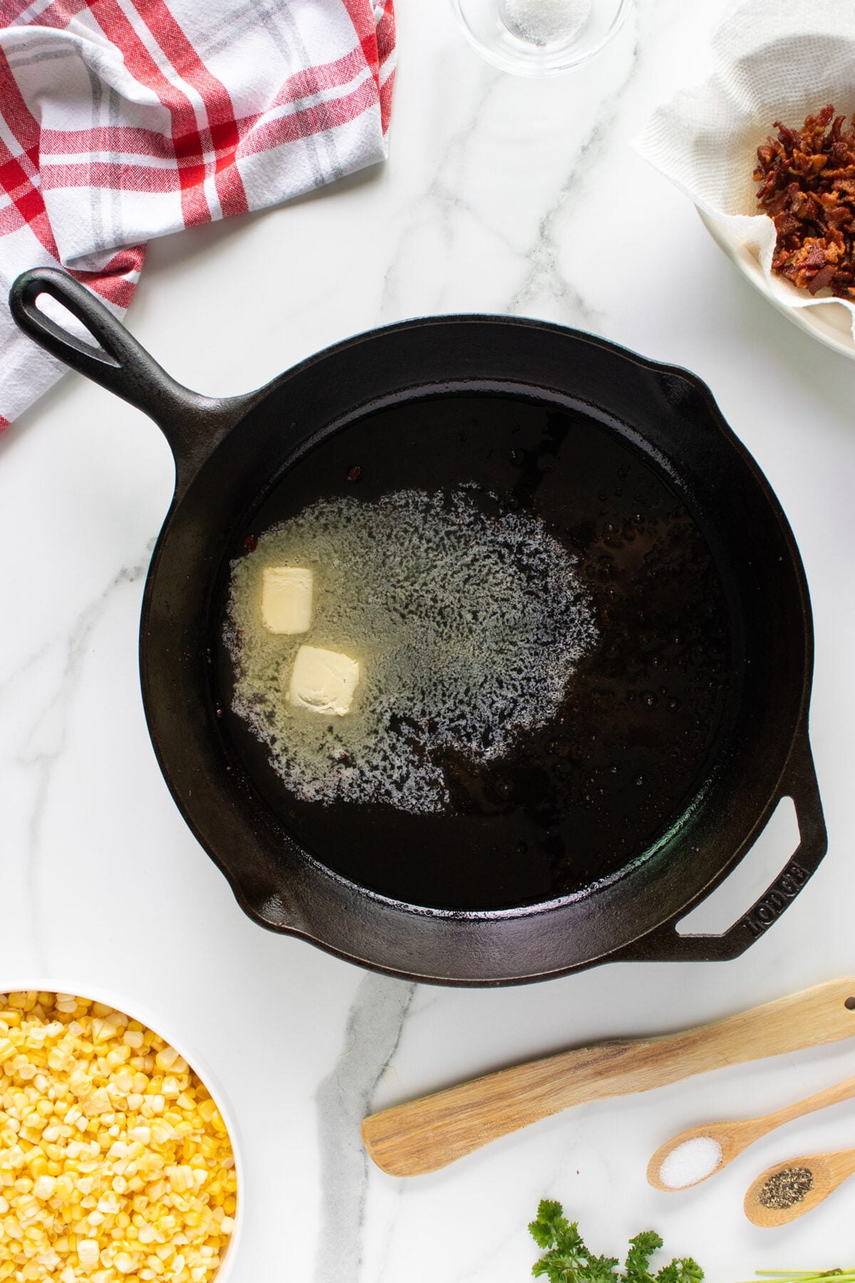 A cast iron skillet with melting butter sits on a white marble countertop, surrounded by a bowl of corn kernels, a wooden spoon, chopped herbs, seasonings, a red and white towel, and a bowl of cooked bacon pieces.