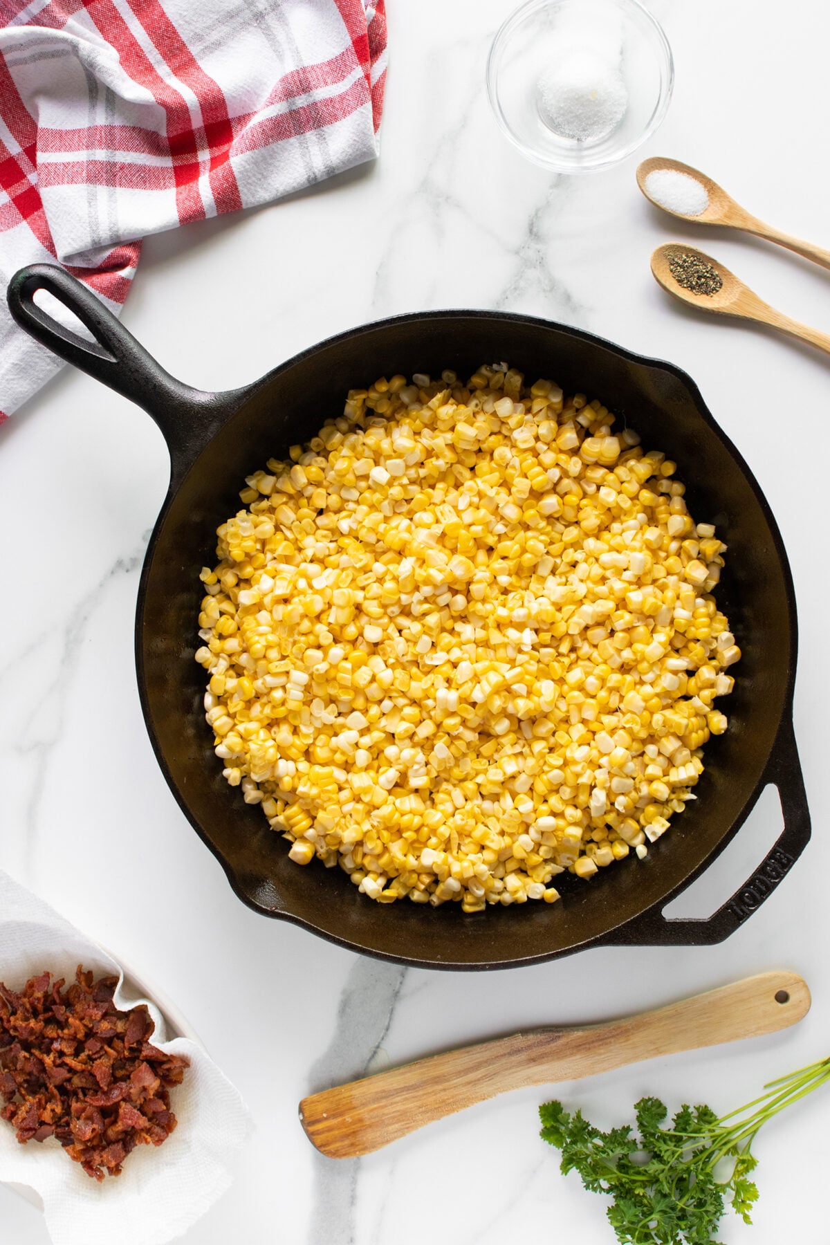 A cast iron skillet filled with kernels of fresh corn sits on a white marble surface. Nearby are wooden spoons with salt and pepper, a glass bowl, chopped bacon on paper towels, a red-striped towel, and fresh parsley.