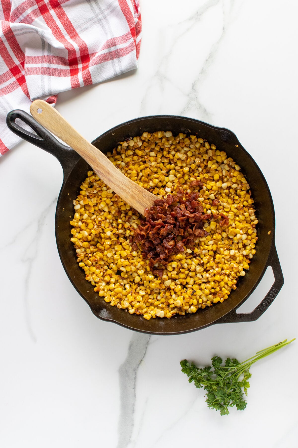 A cast iron skillet filled with cooked corn and topped with crumbled bacon, stirred with a wooden spatula. A red and white kitchen towel and a sprig of parsley are nearby on a white marble surface.