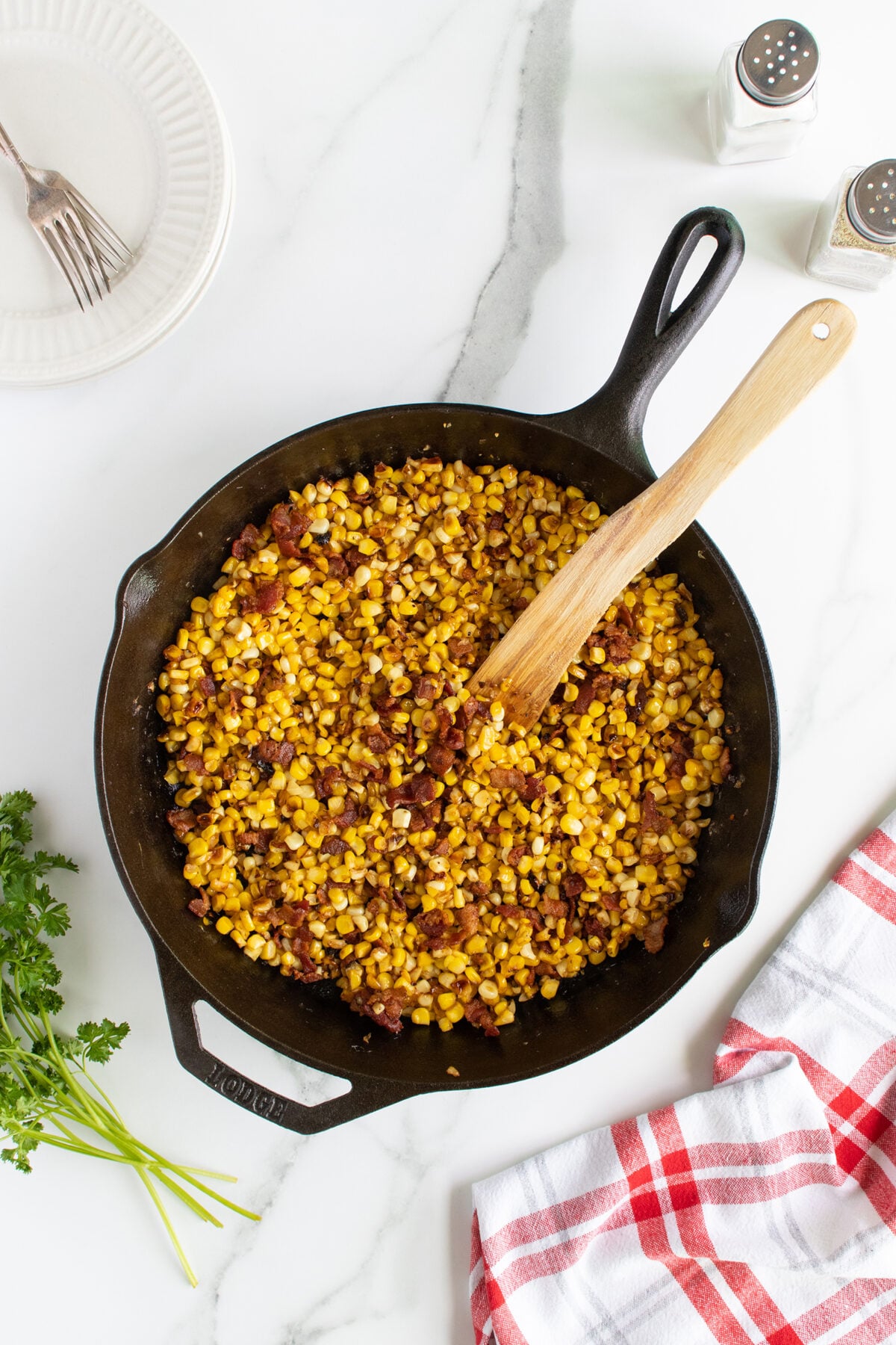 A cast iron skillet filled with sautéed corn and bacon sits on a marble countertop. A wooden spoon rests in the skillet. Nearby are a white plate with a fork, salt and pepper shakers, fresh parsley, and a red plaid towel.