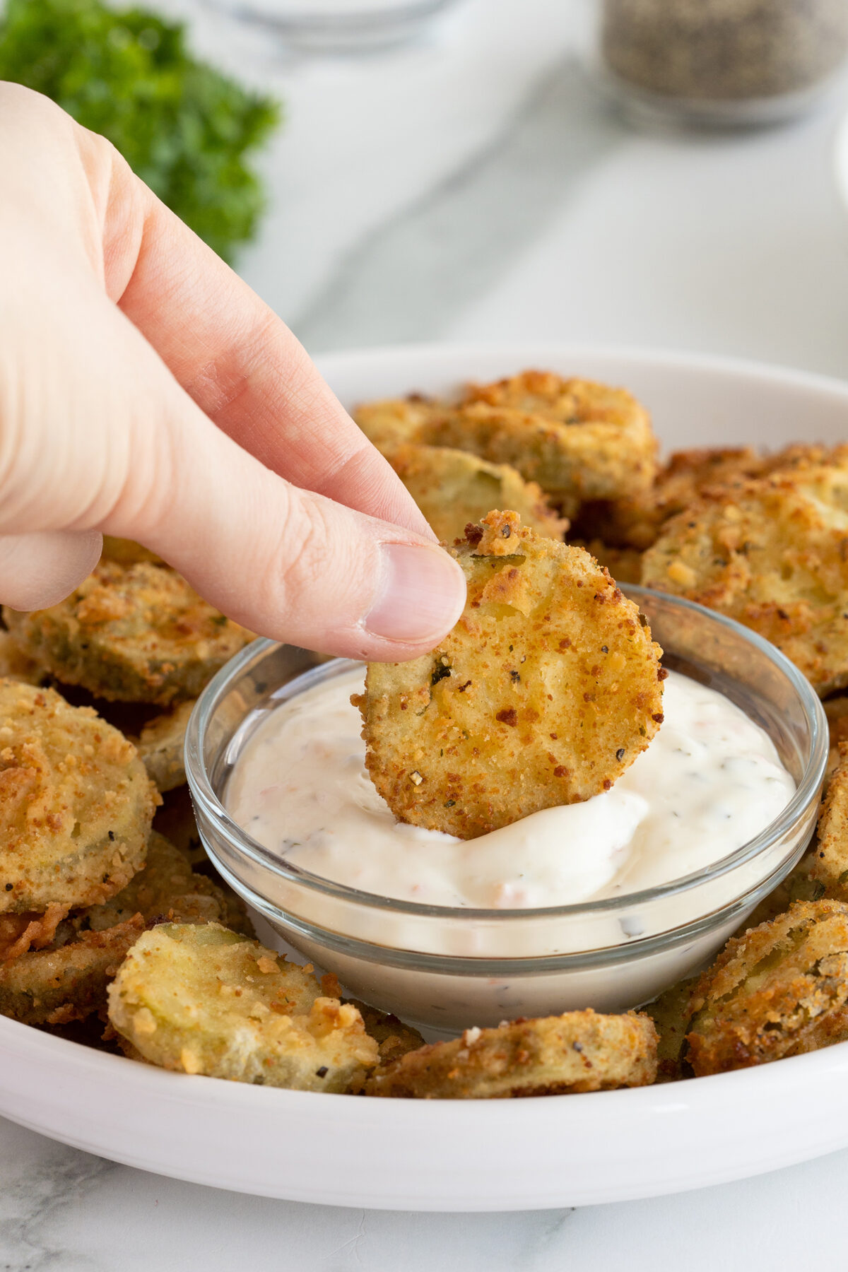 A hand dips a fried pickle slice into a bowl of creamy dipping sauce, with more fried pickles arranged on a plate underneath.