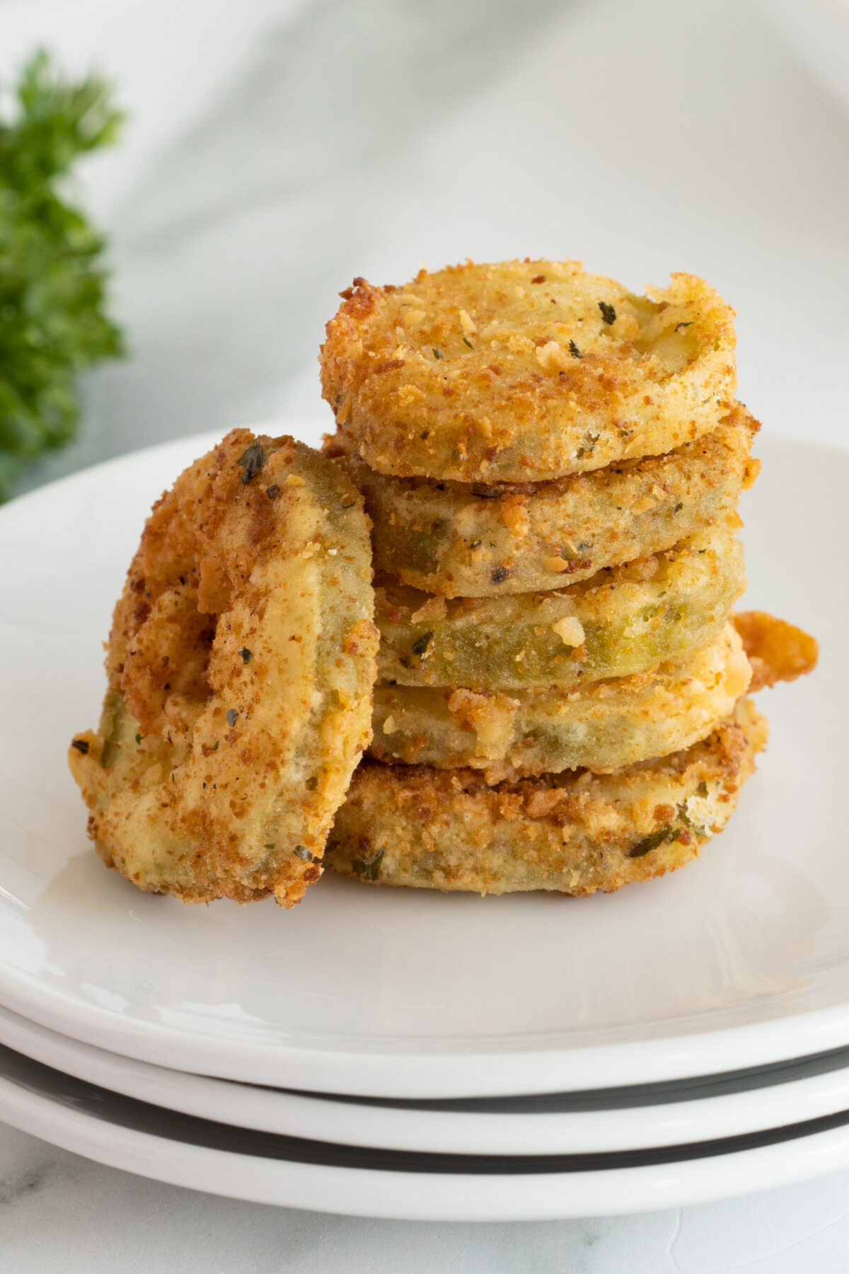 A stack of crispy, golden-brown fried green tomato slices sits on a white plate with a blurred green garnish in the background.