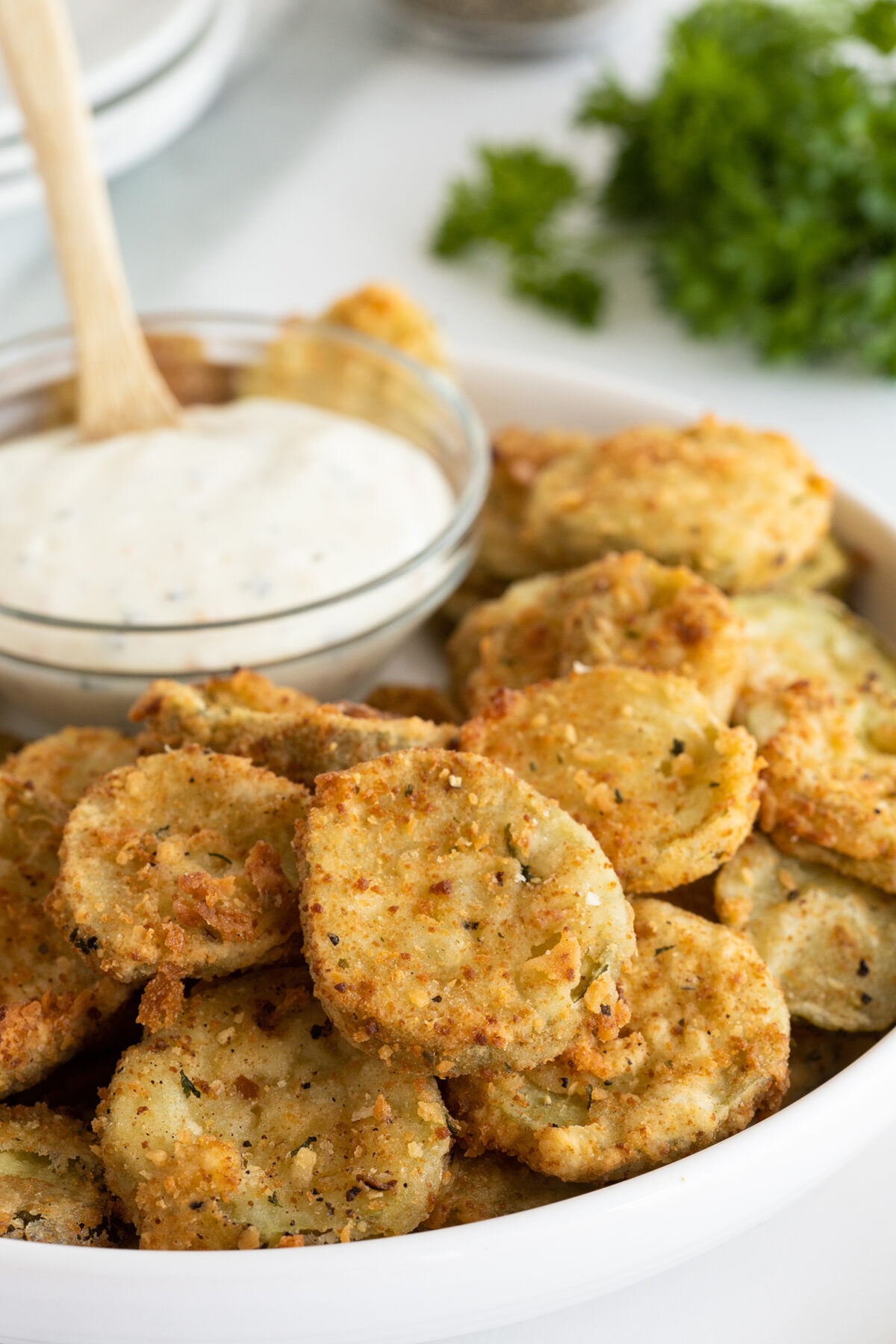 A plate of crispy fried pickles sits next to a glass bowl of creamy dipping sauce, with fresh parsley in the background.