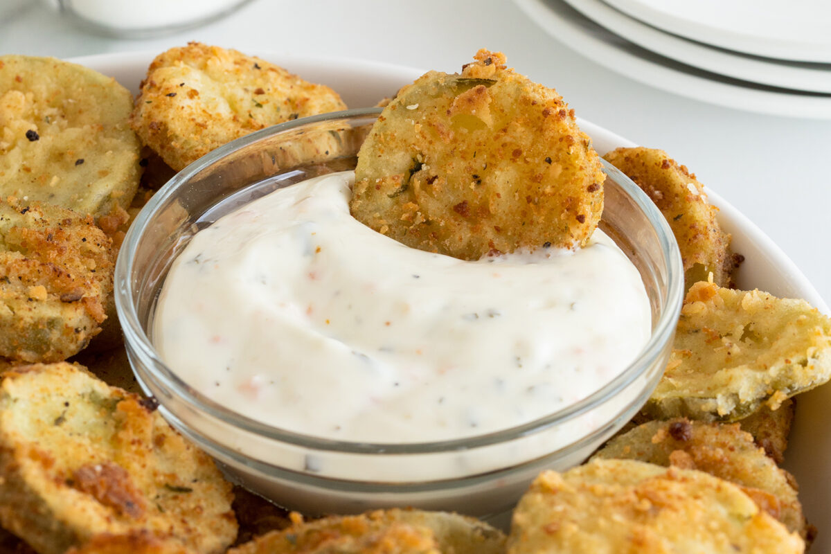 A bowl of creamy dipping sauce is surrounded by crispy fried pickle slices, with one pickle slice partially dipped in the sauce. White plates are visible in the background.