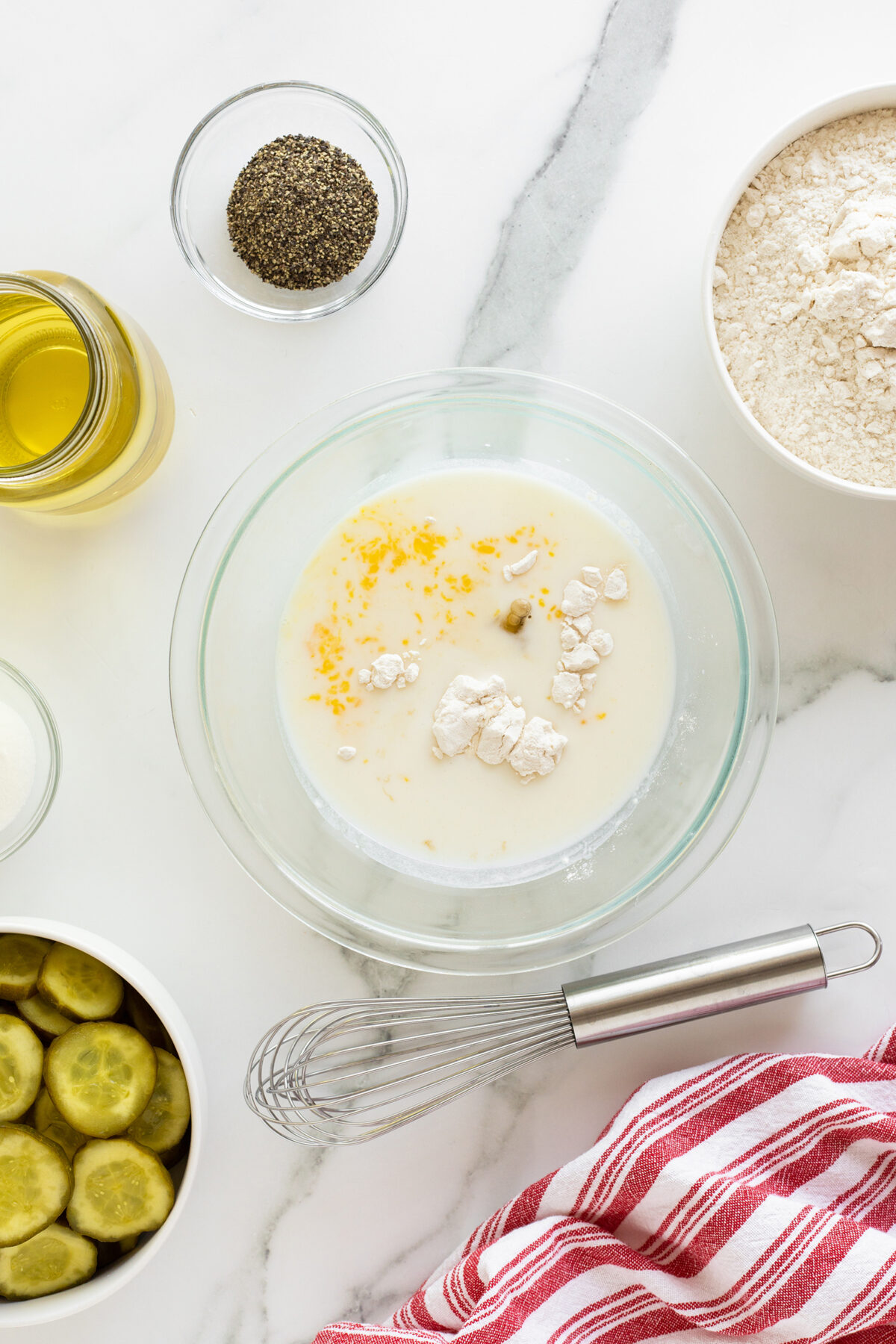A glass bowl with milk, flour, and lemon zest sits on a marble counter surrounded by bowls of flour, black pepper, sliced pickles, oil, a whisk, and a red-striped towel.