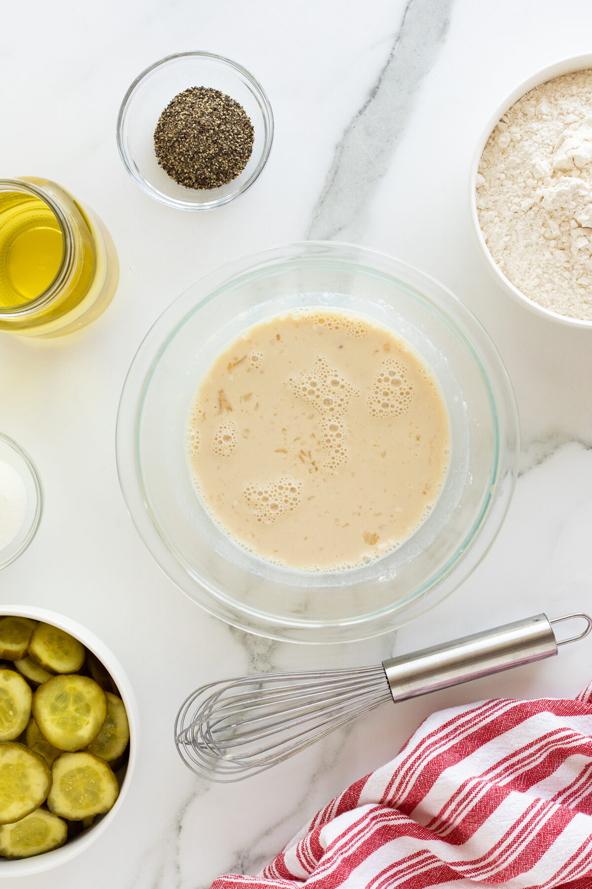 A glass bowl of foamy liquid sits on a marble surface, surrounded by a whisk, a red-striped towel, flour, black pepper, oil, sugar, and a bowl of sliced pickles.