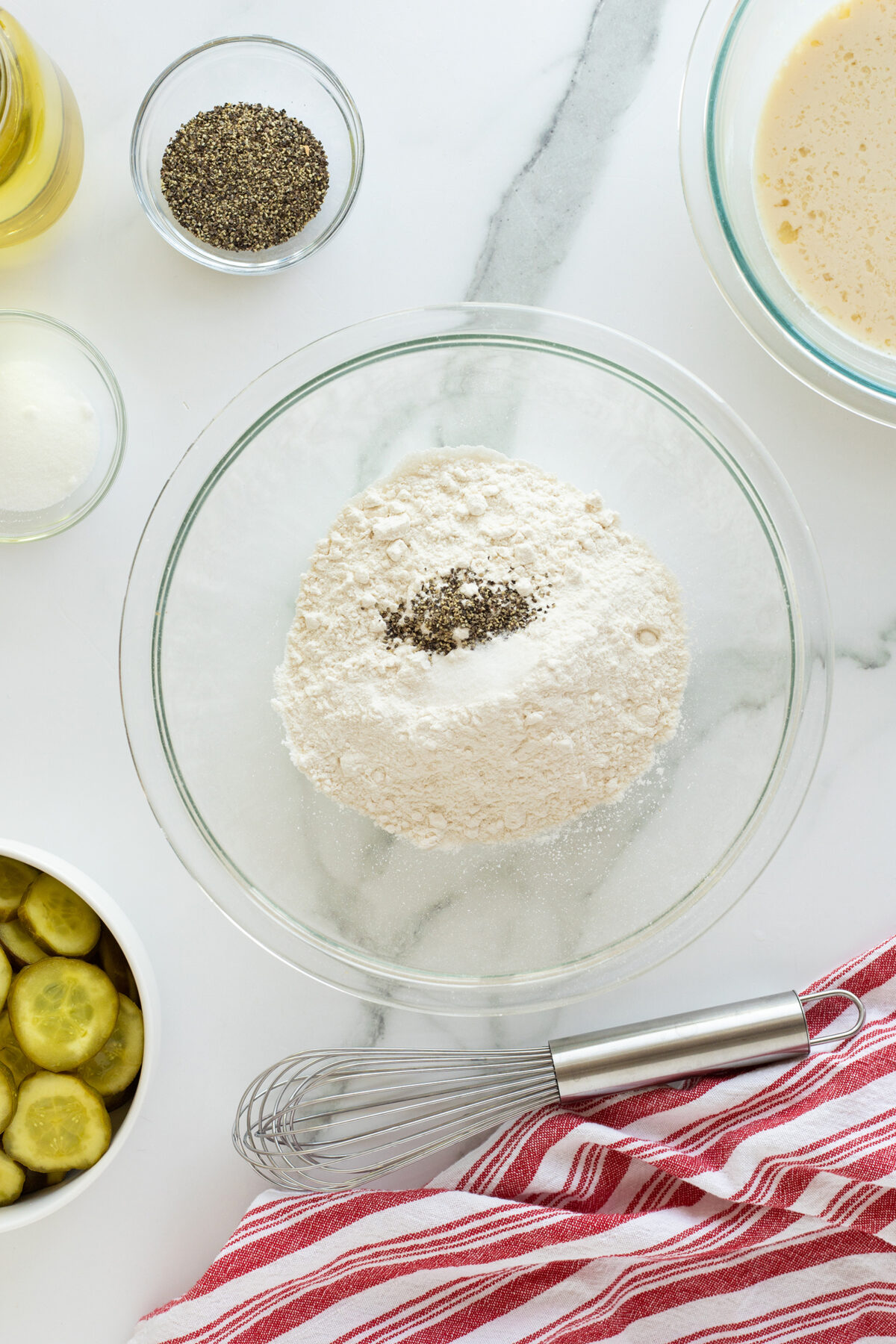 A glass bowl with flour and pepper in the center, surrounded by a whisk, a red striped towel, sliced pickles, a small bowl of pepper, a bowl of liquid, and an oil bottle on a white marble surface.