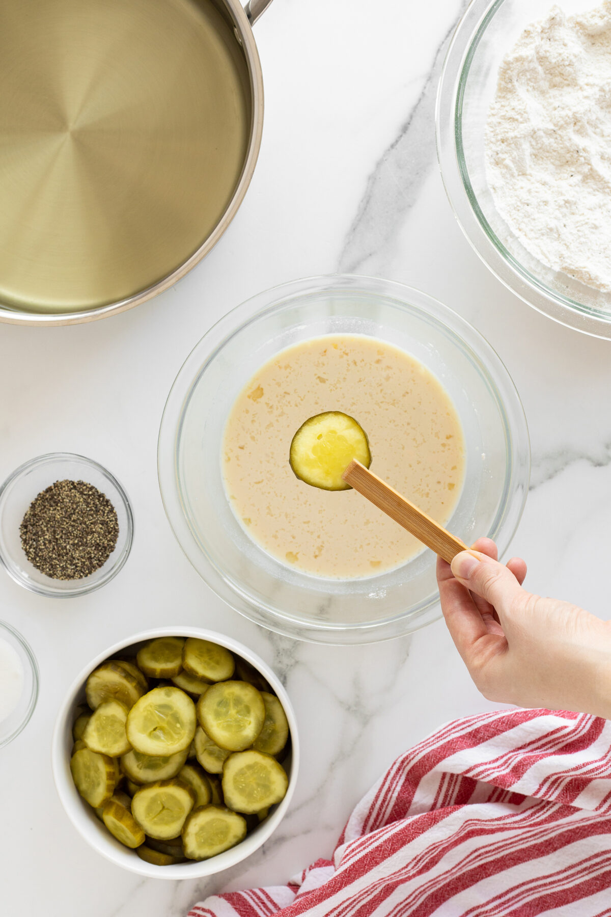 A hand dips a pickle slice into a bowl of batter. Nearby are bowls of flour, sliced pickles, black pepper, and an empty saucepan on a marble countertop with a red-striped kitchen towel.
