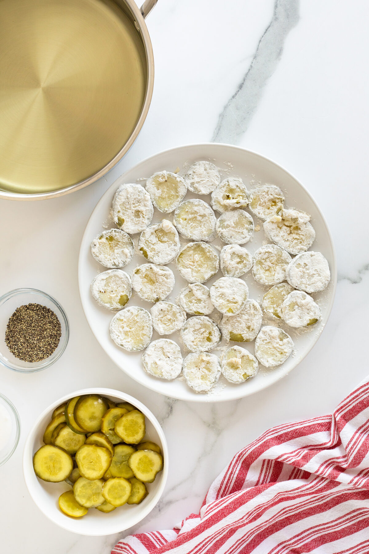 A plate of flour-coated pickle slices sits beside a bowl of sliced pickles, a bowl of black pepper, a pot, and a red-striped kitchen towel on a white marble countertop.