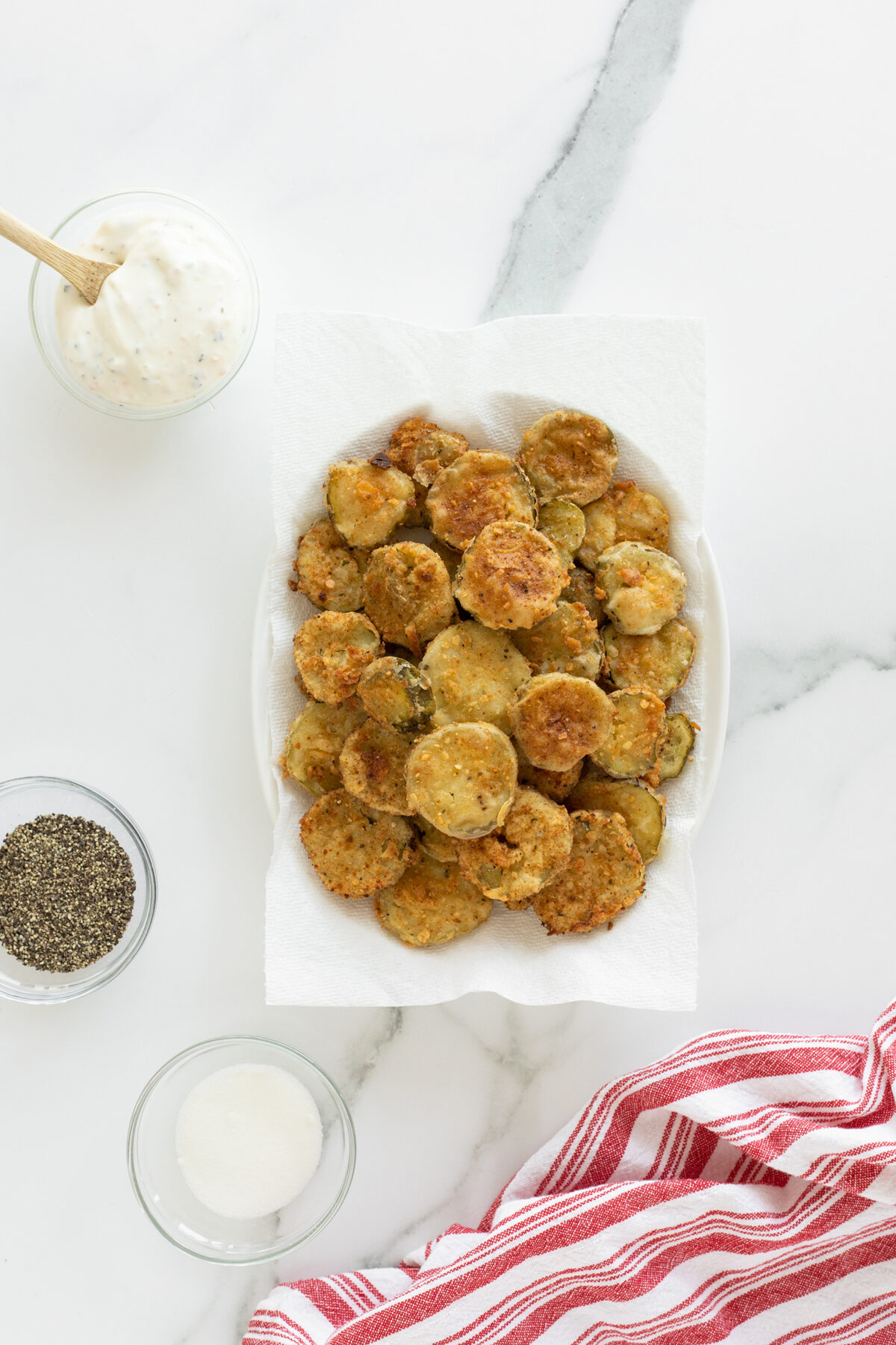 A white plate of fried pickles sits on a paper towel, surrounded by small bowls of ranch dip, black pepper, and salt. A red and white striped cloth lies nearby on a marble surface.