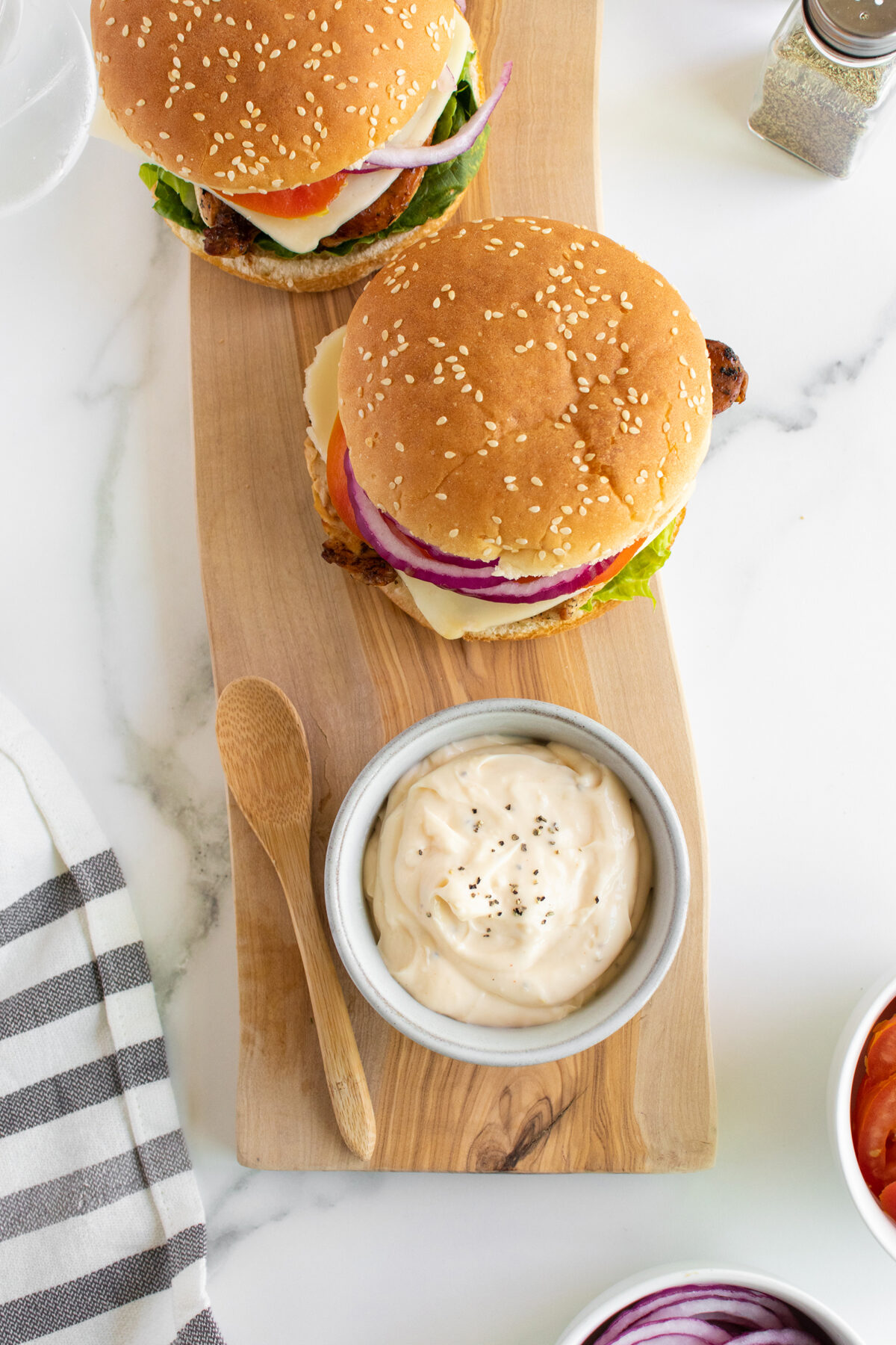 Two cheeseburgers with lettuce, tomato, onion, and sauce on a wooden board next to a bowl of mayonnaise sprinkled with black pepper, a wooden spoon, and a bowl of sliced onions.