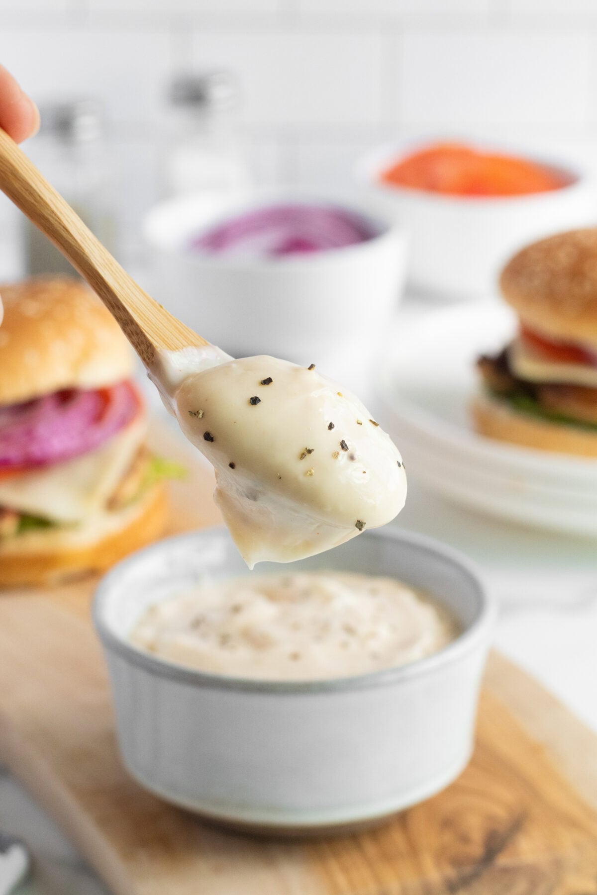 A wooden spoon holds a creamy peppered sauce above a small bowl, with burgers and bowls of toppings like sliced onion and tomato in the blurred background.