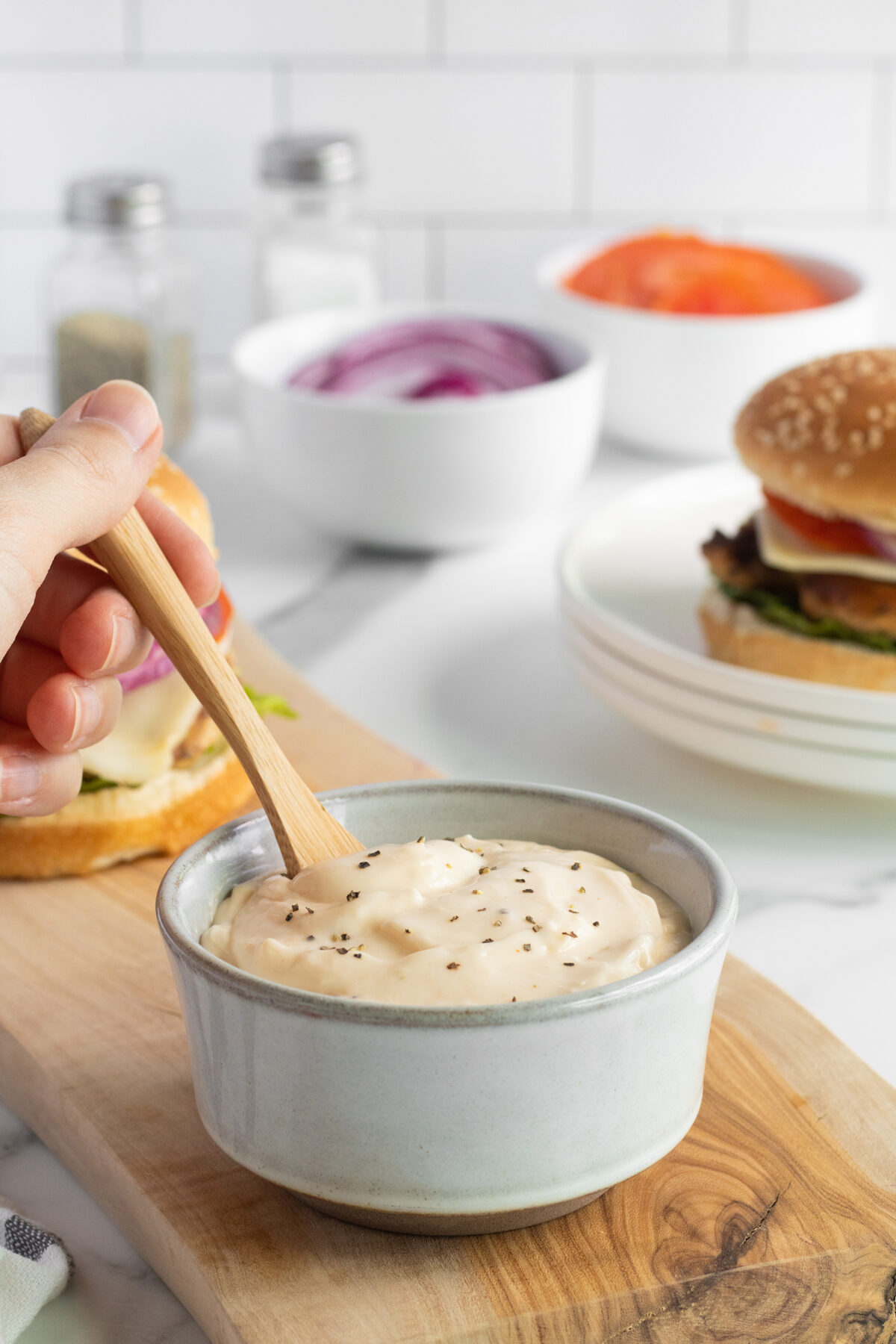 A hand stirs creamy sauce topped with black pepper in a small bowl on a wooden board. In the background are a burger, sliced onions, tomatoes, and condiment shakers.