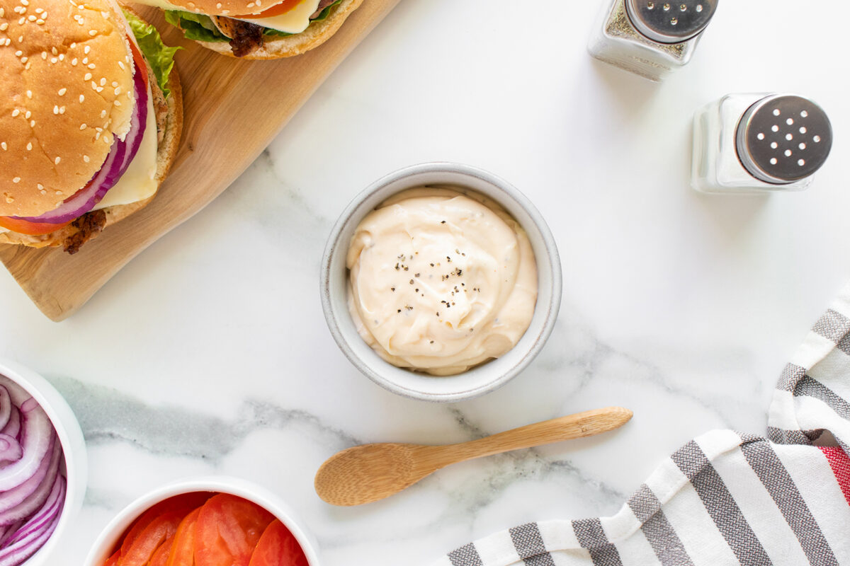 A bowl of creamy sauce with black pepper, a wooden spoon, a hamburger, sliced onions, sliced tomatoes, salt and pepper shakers, and a striped cloth on a marble surface.