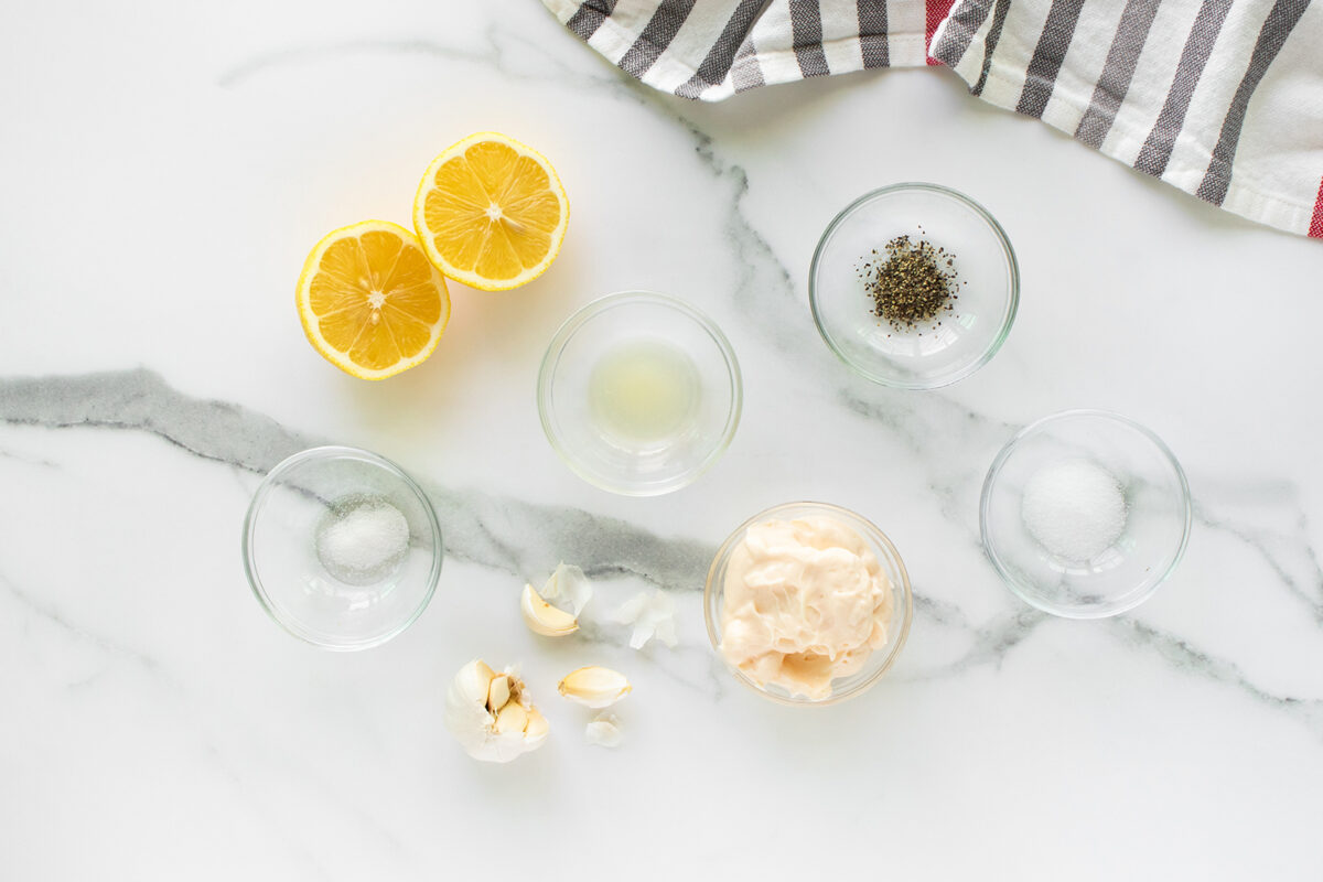 A marble counter with halved lemons, small bowls of pepper, salt, mayonnaise, lemon juice, and garlic cloves. A striped cloth is at the top right corner.