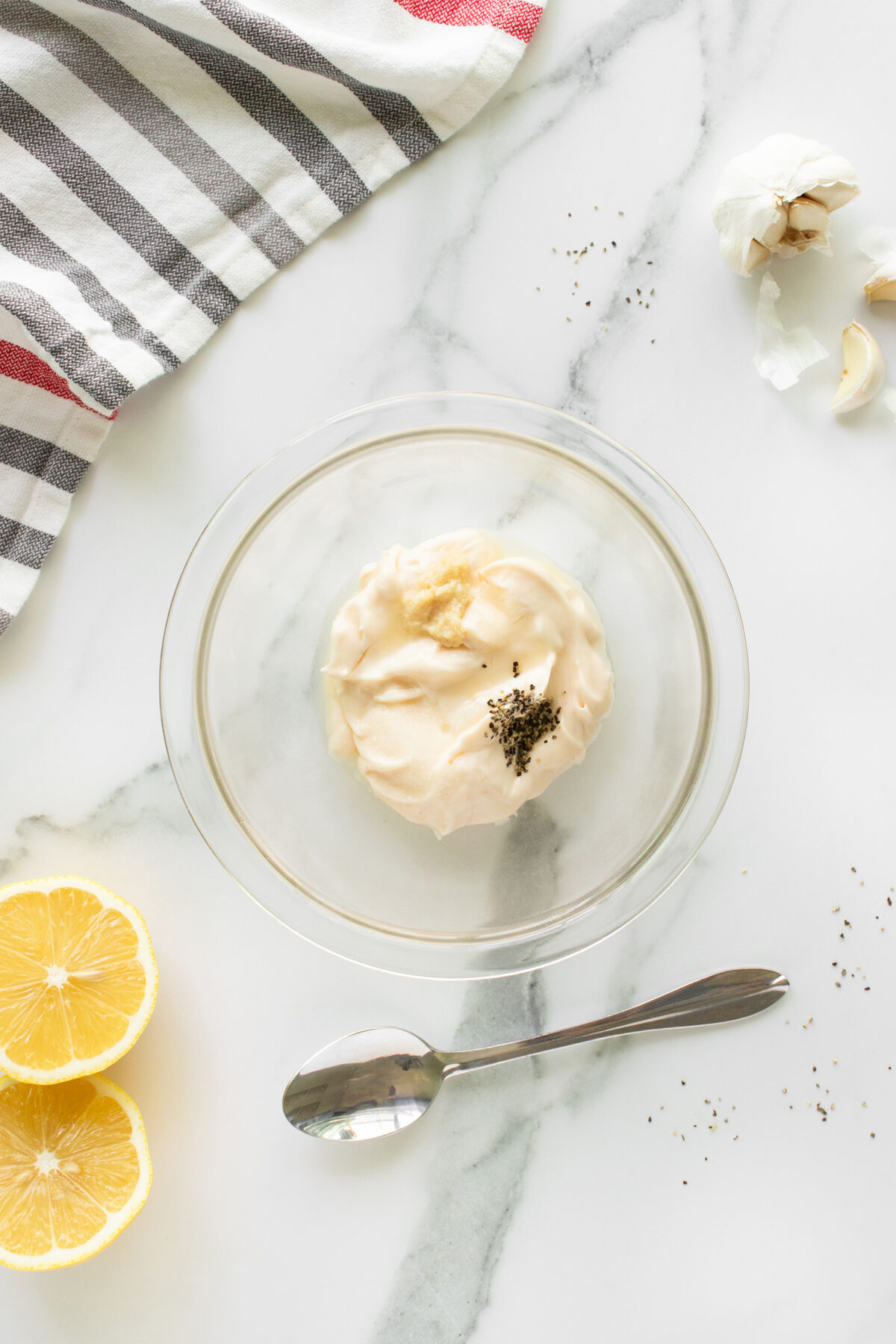 A glass bowl with mayonnaise, minced garlic, and black pepper sits on a marble surface. Nearby are a spoon, lemon halves, garlic cloves, scattered pepper, and a striped kitchen towel.