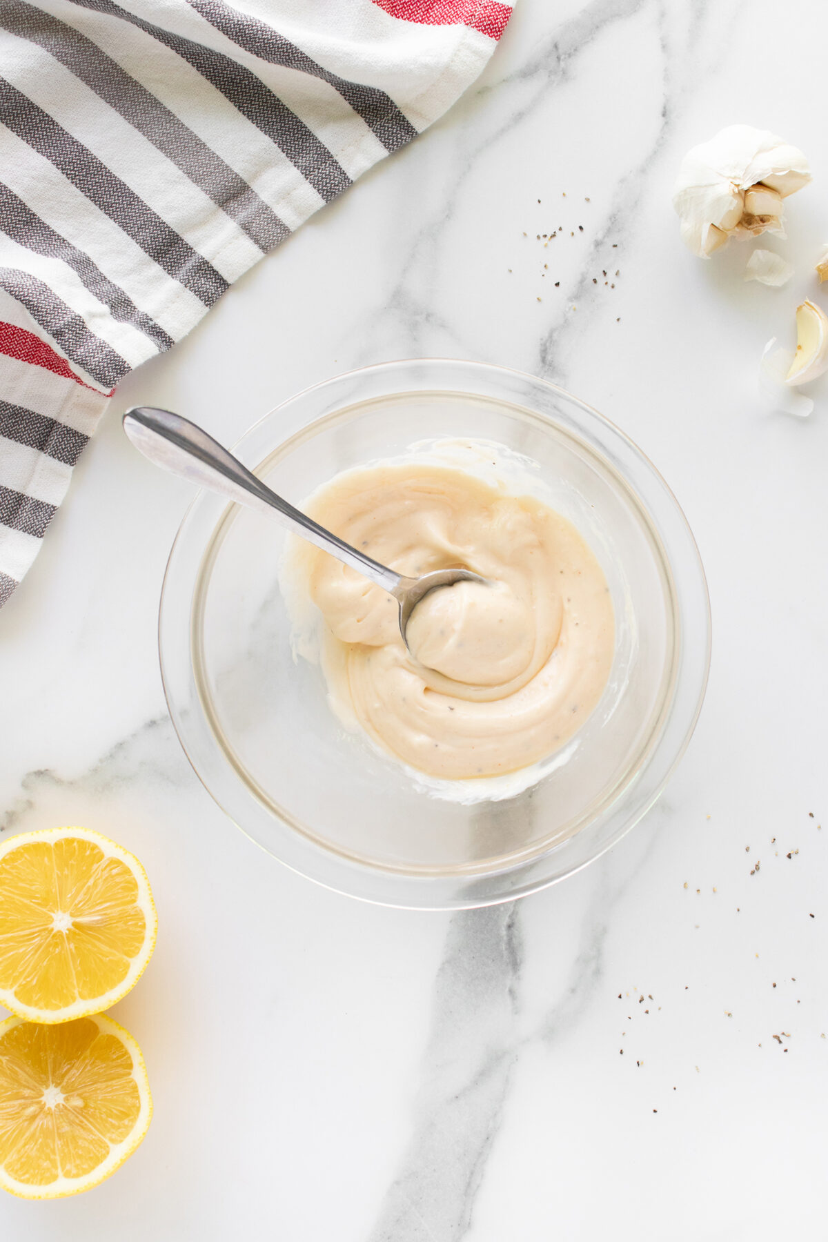 A glass bowl with creamy dressing and a spoon on a marble surface, surrounded by lemon halves, garlic, scattered pepper, and a striped kitchen towel.