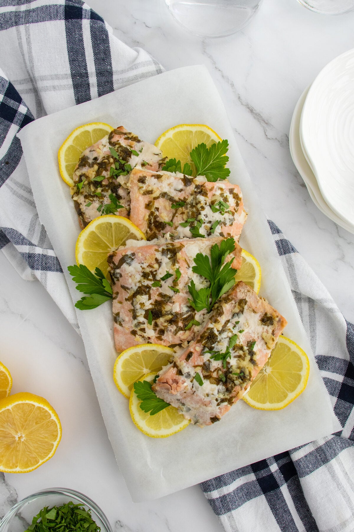 Three pieces of herb-crusted baked fish are arranged on parchment paper with lemon slices and parsley garnish, placed on a white surface near a blue plaid napkin and stacked white plates.