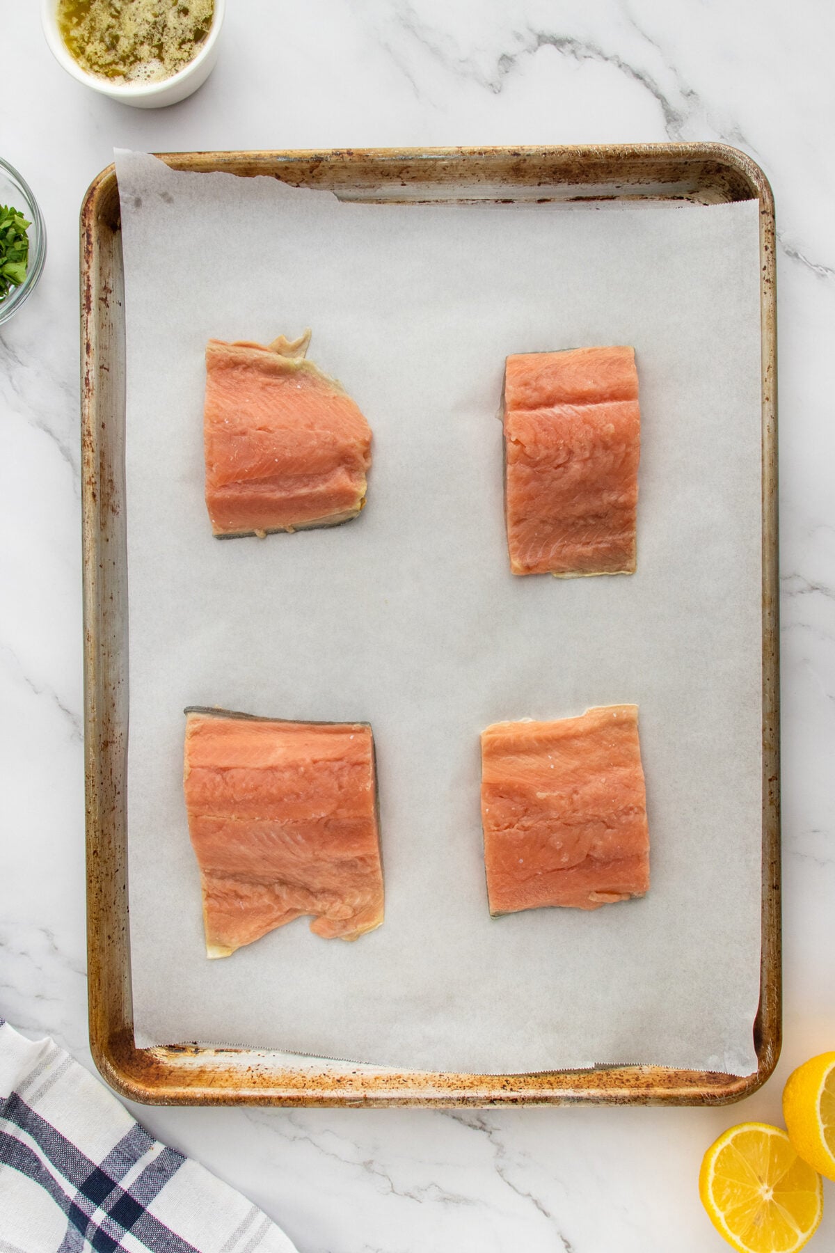 Four raw salmon fillets are placed on a parchment-lined baking sheet. Nearby are a bowl of seasoning, chopped herbs, and lemon halves. The scene is set on a marble countertop with a checkered napkin.