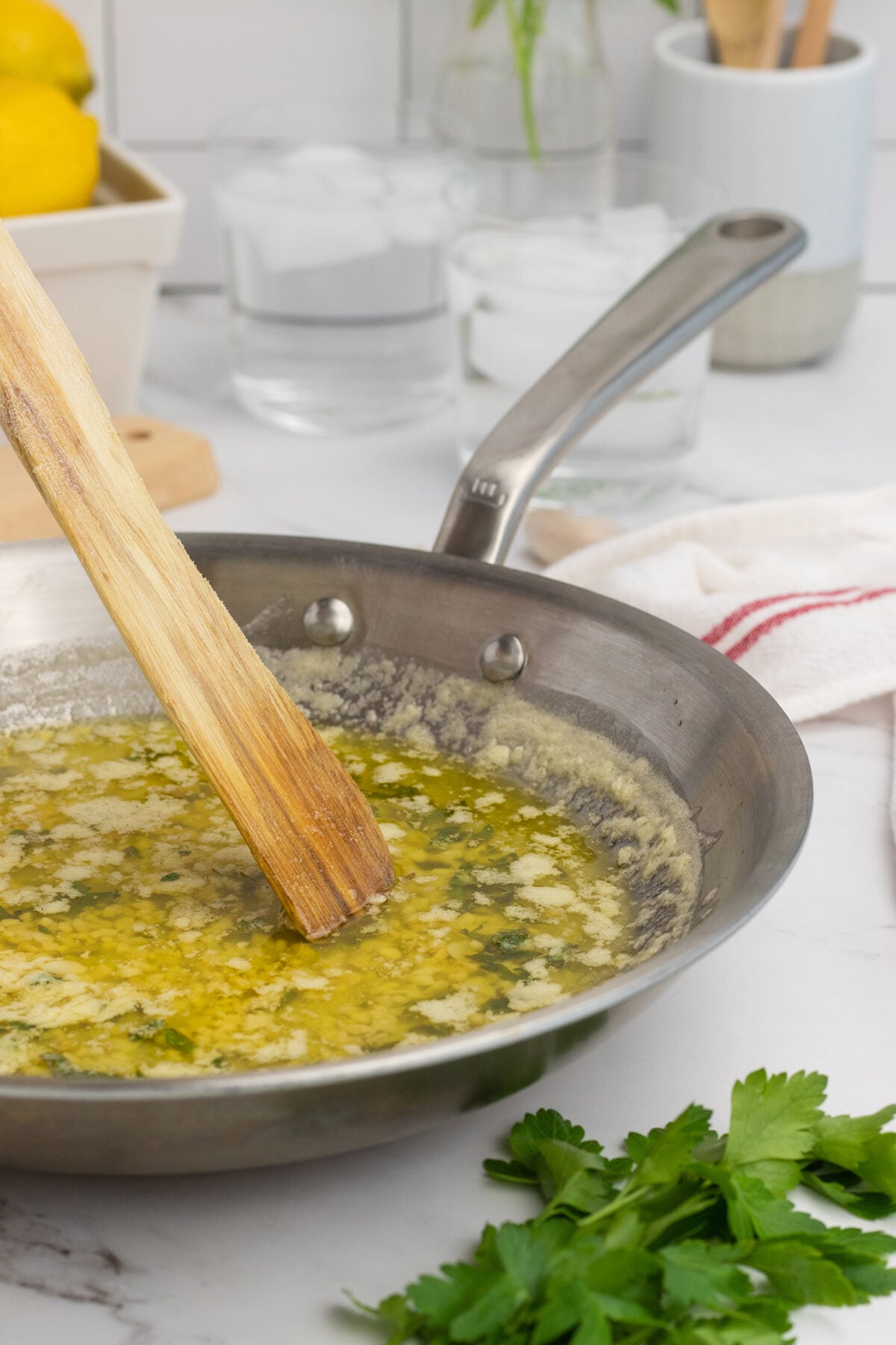 A wooden spatula stirs melted butter with herbs in a stainless steel skillet. Fresh parsley sits nearby on a white countertop, with kitchen items and glasses of ice water in the background.
