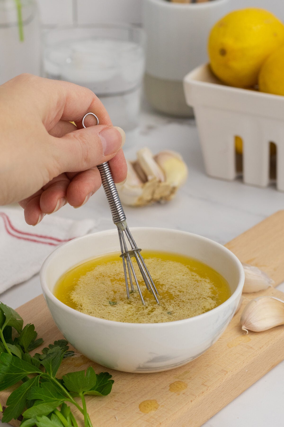 A hand uses a small whisk to mix oil and seasoning in a white bowl on a kitchen counter, with garlic, parsley, lemons, and a glass of water in the background.