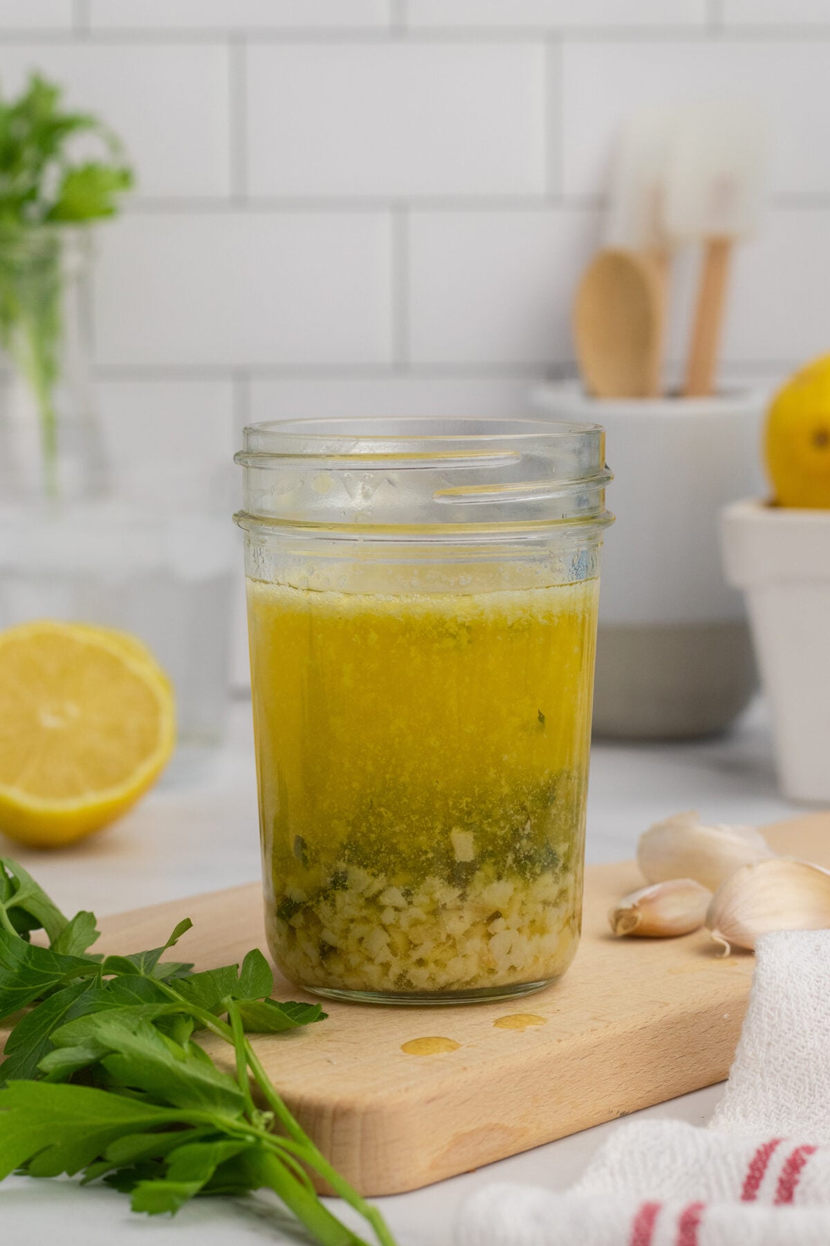 A glass jar filled with homemade salad dressing sits on a wooden board. Chopped herbs and garlic are visible at the bottom, with oil and vinegar layered on top. Parsley, garlic, a lemon half, and kitchen utensils are in the background.
