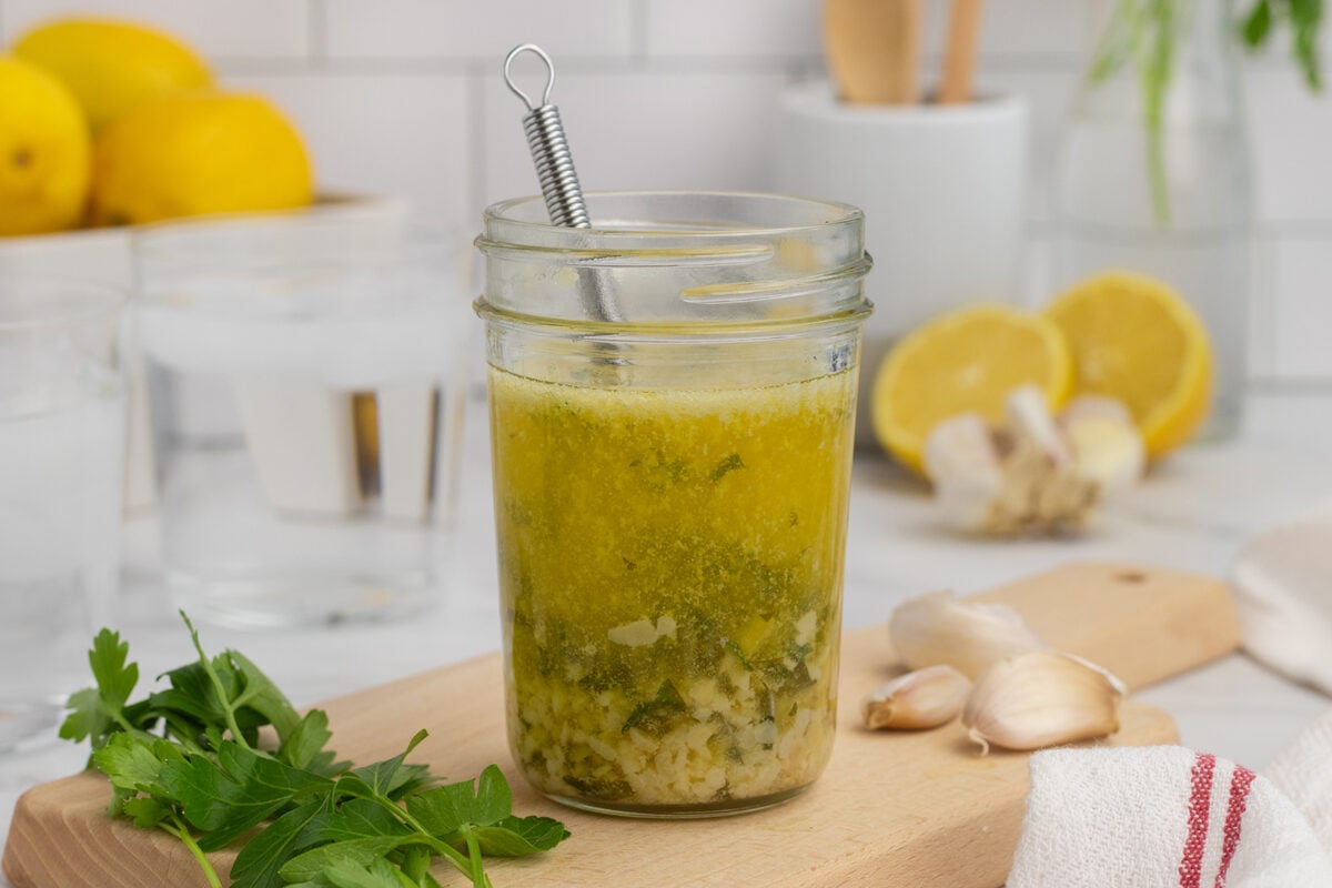 A glass jar of homemade vinaigrette with herbs and diced garlic sits on a wooden board, surrounded by fresh parsley, garlic cloves, and sliced lemons. A small whisk is inside the jar, and kitchen items are blurred in the background.