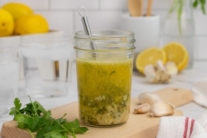 A glass jar of homemade vinaigrette with herbs and diced garlic sits on a wooden board, surrounded by fresh parsley, garlic cloves, and sliced lemons. A small whisk is inside the jar, and kitchen items are blurred in the background.