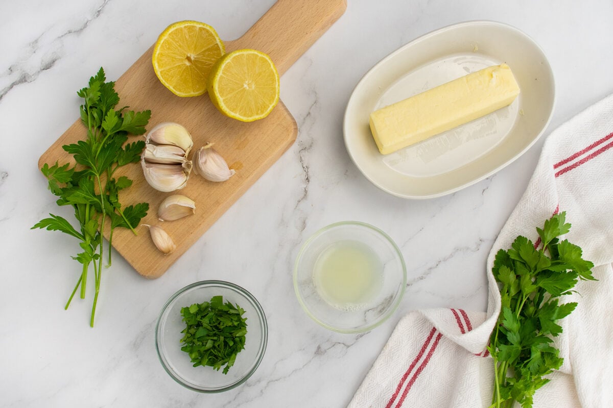 A stick of butter, garlic cloves, parsley, and halved lemon on a white marble surface with a cutting board, small bowl of lemon juice, and a white towel with red stripes.