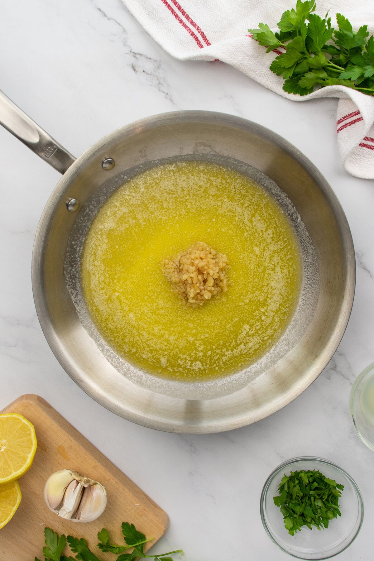 A stainless steel pan with melted butter and minced garlic in the center, surrounded by fresh parsley, lemon halves, garlic cloves, and chopped herbs on a white marble surface.