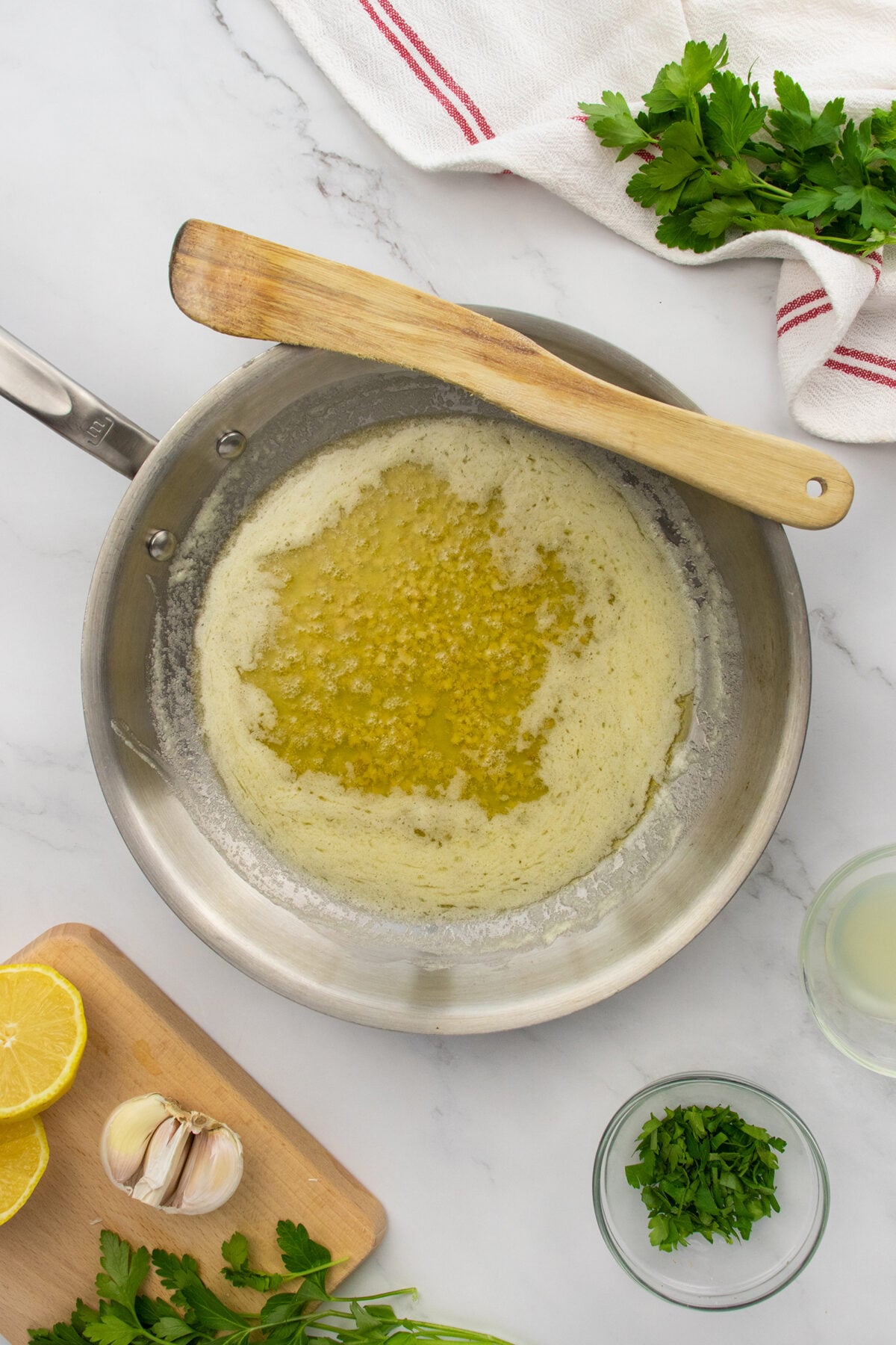 Overhead view of a skillet with melted butter on a marble surface, surrounded by fresh parsley, lemon halves, garlic cloves, and a wooden spatula resting on the pan. A kitchen towel is in the upper corner.