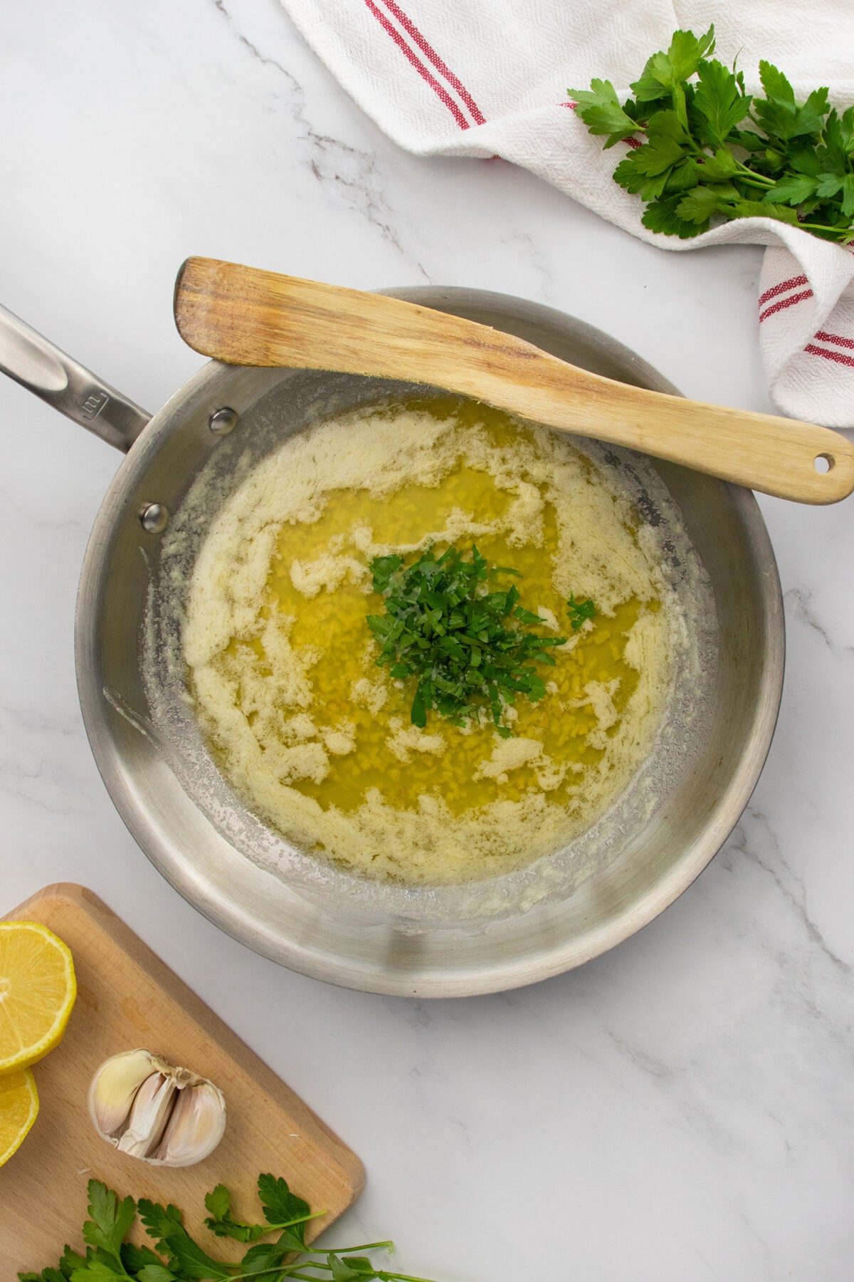A frying pan with melted butter and chopped parsley in the center, resting on a marble counter. Nearby are a wooden spatula, a cutting board with lemon, garlic, and parsley, and a white towel with red stripes.