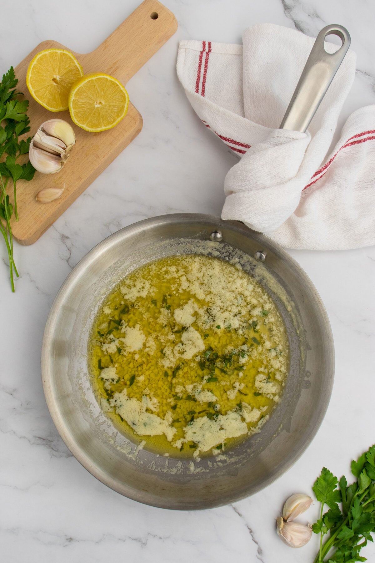 A metal skillet with melted butter and herbs sits on a marble countertop, surrounded by fresh parsley, garlic, a halved lemon on a wooden board, and a white kitchen towel with a red stripe.
