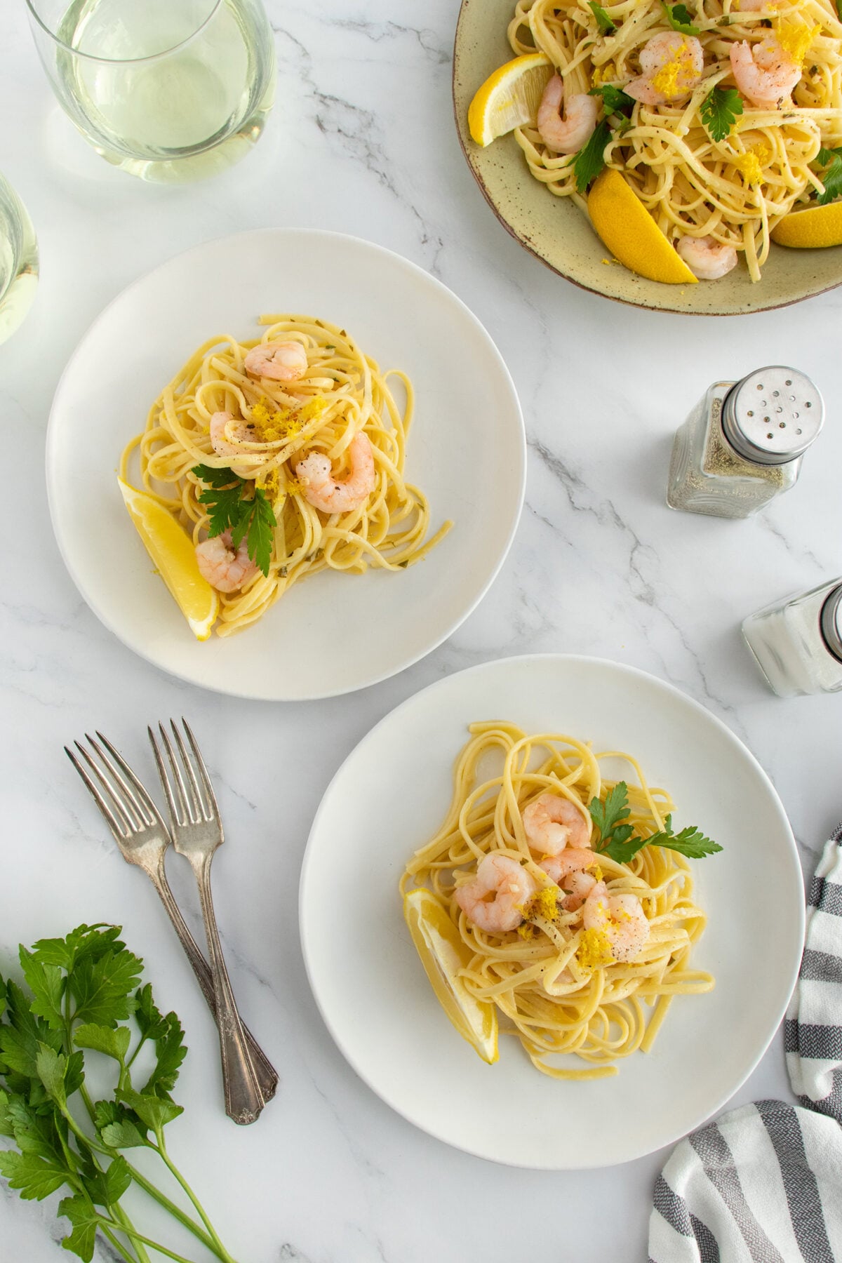 Two white plates with shrimp scampi pasta garnished with parsley and lemon wedges sit on a marble table; a serving platter, salt shaker, wine glasses, fresh parsley, and forks are beside them.