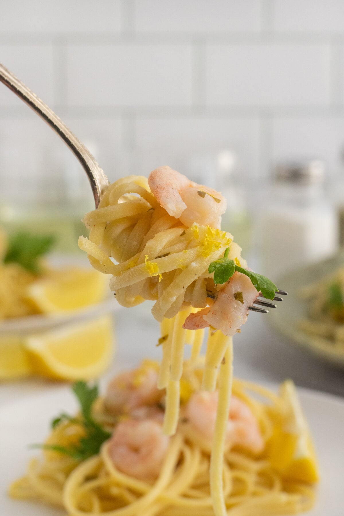 A close-up of a fork twirling spaghetti with shrimp, garnished with parsley and lemon zest, above a plate of pasta. The background shows a blurred table setting and white tiled wall.