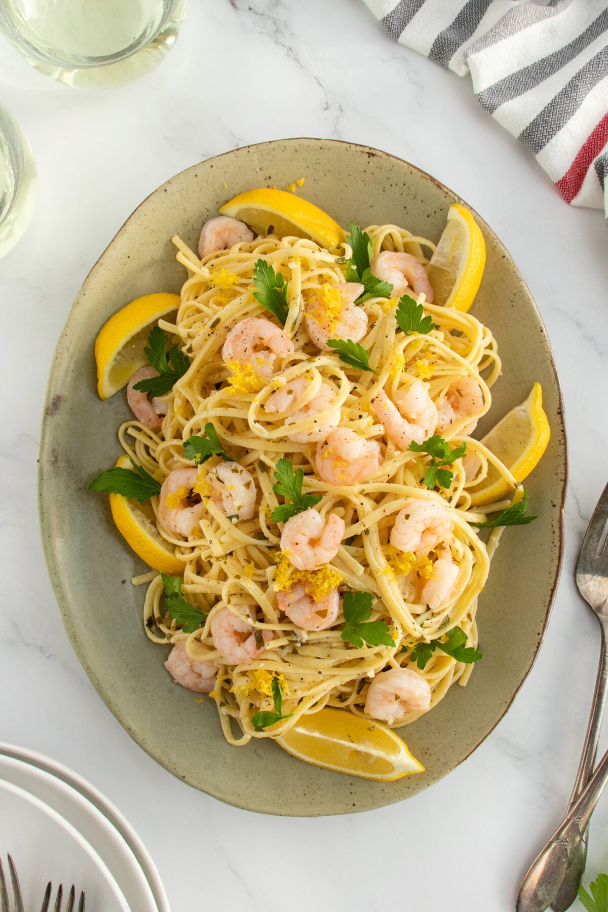 A plate of pasta topped with shrimp, fresh parsley, and lemon zest, garnished with lemon wedges. The plate sits on a marble surface with a fork, knife, and striped napkin nearby.